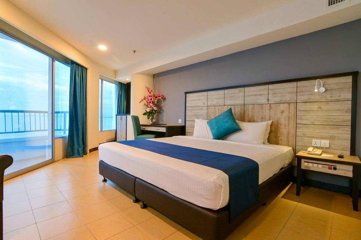 A hotel room with a large bed, a nightstand, a desk, and a view of the sea.