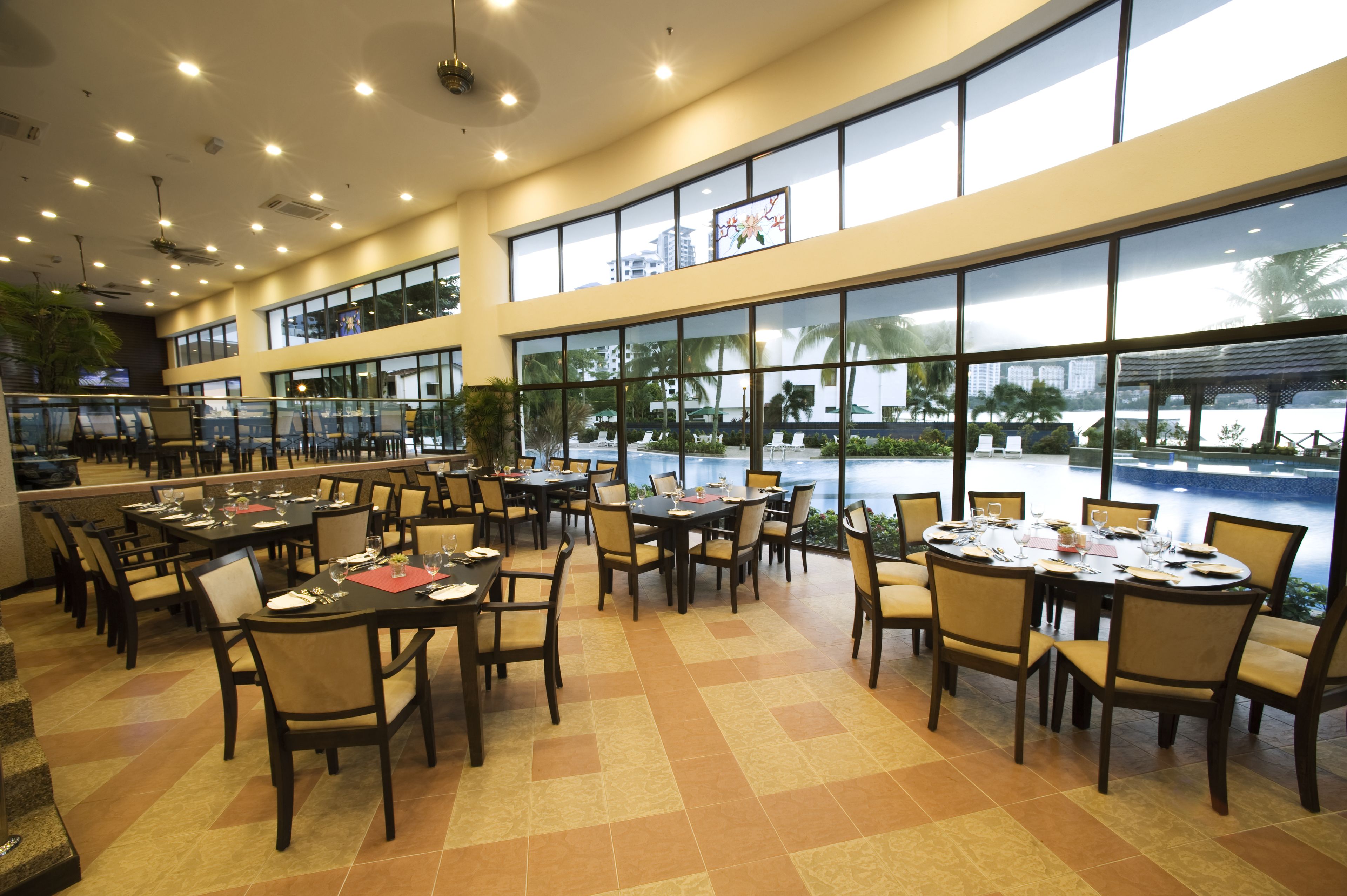 Un comedor amplio con varias mesas y sillas, ventanas grandes que ofrecen vistas a una piscina y palmeras, y un diseño de techo moderno con iluminación empotrada.