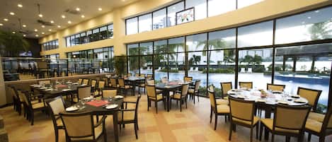 Uma ampla sala de jantar com várias mesas e cadeiras, grandes janelas com vista para uma piscina e palmeiras, e um design de teto moderno com iluminação embutida.