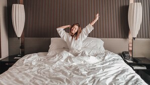 Habitación de hotel con cama, mesita de noche, lámpara y una pared con persianas verticales.