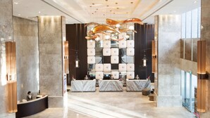 Un moderno vestíbulo de hotel con mostradores de recepción de mármol, un candelabro central y amplias ventanas.