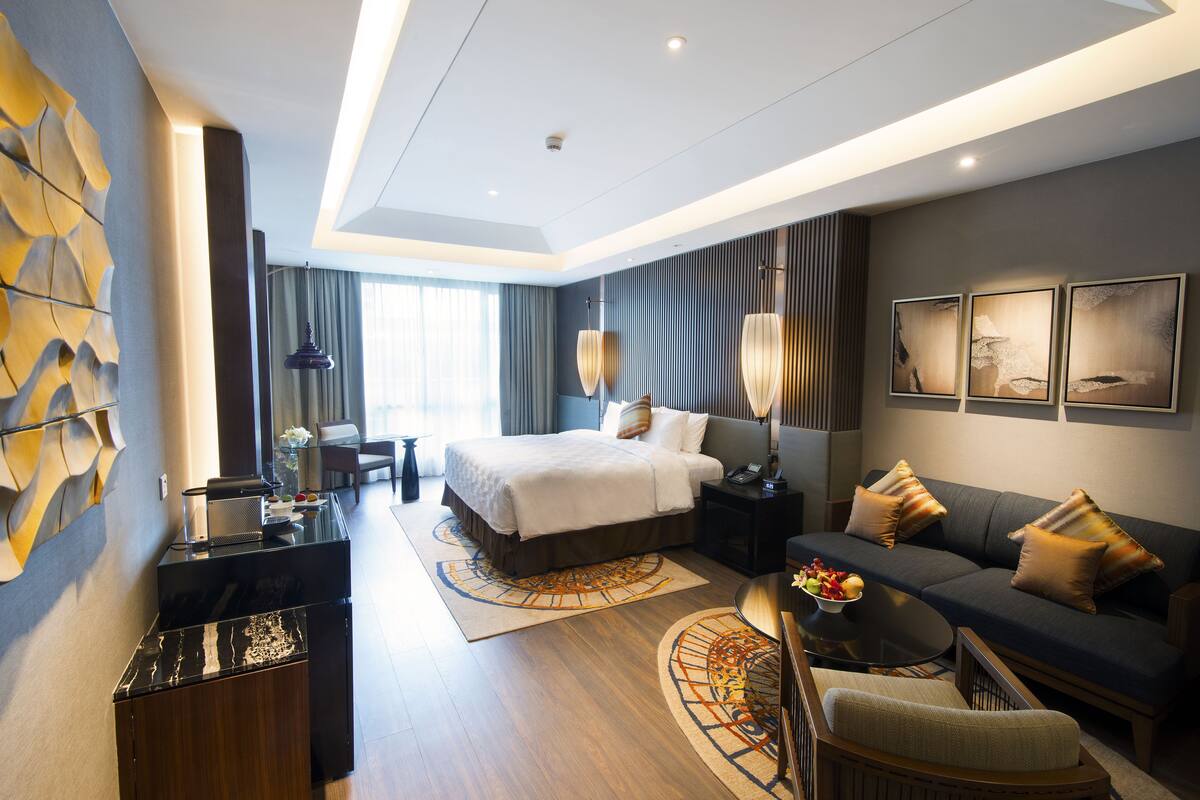 A modern hotel room with a large bed, a sofa, a desk, and a dining area.