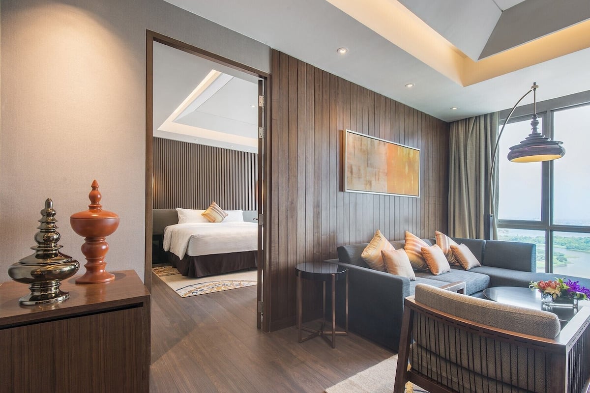 A modern hotel room with a bed, a sofa, a desk, and a large window offering a view.