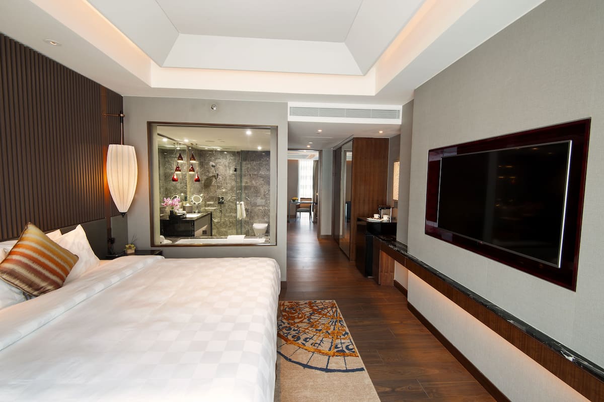 A modern hotel room with a large bed, a TV, a bathroom with a glass shower, and a hallway leading to other rooms.