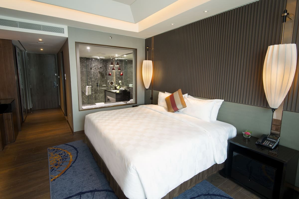 A modern hotel room with a large bed, a bedside table with a lamp, a mirror, and a view of the bathroom.