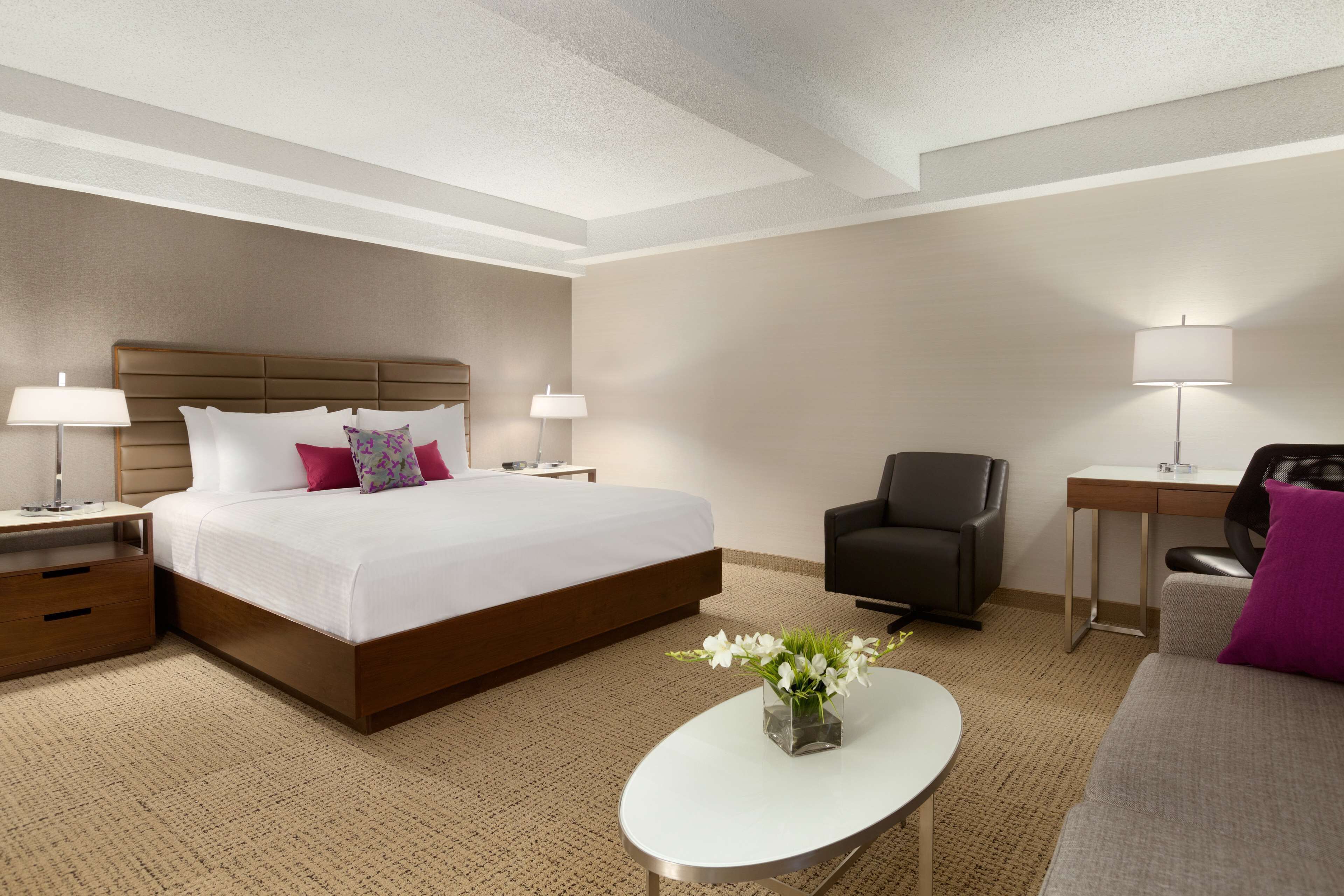 A hotel room with a large bed, two bedside tables with lamps, a desk, a chair, and a small table with a vase of flowers.