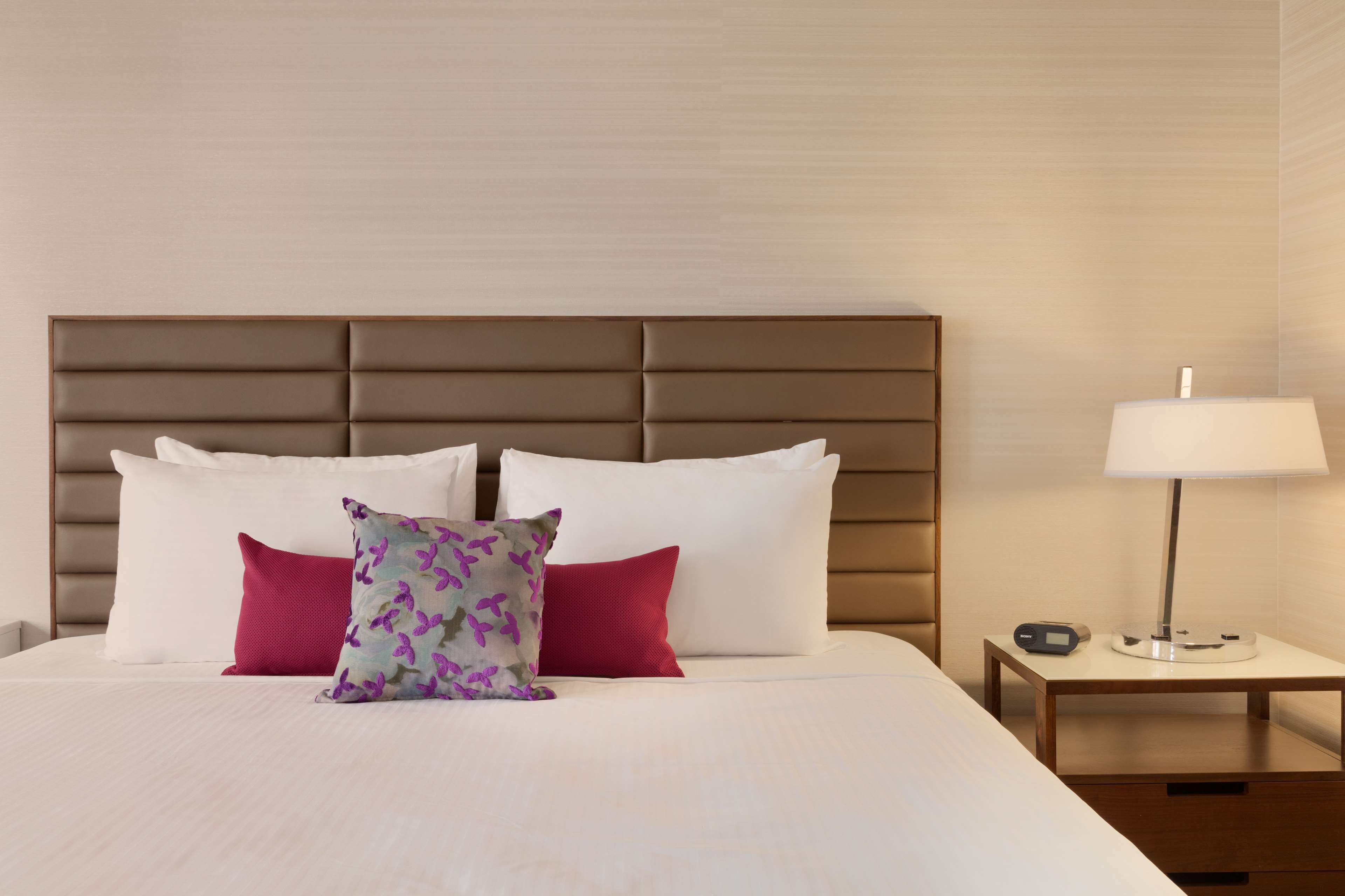 A neatly made bed with white and red pillows, a patterned pillow, and a bedside table with a lamp and a clock.