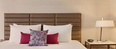 A neatly made bed with white and red pillows, a patterned pillow, and a bedside table with a lamp and a clock.