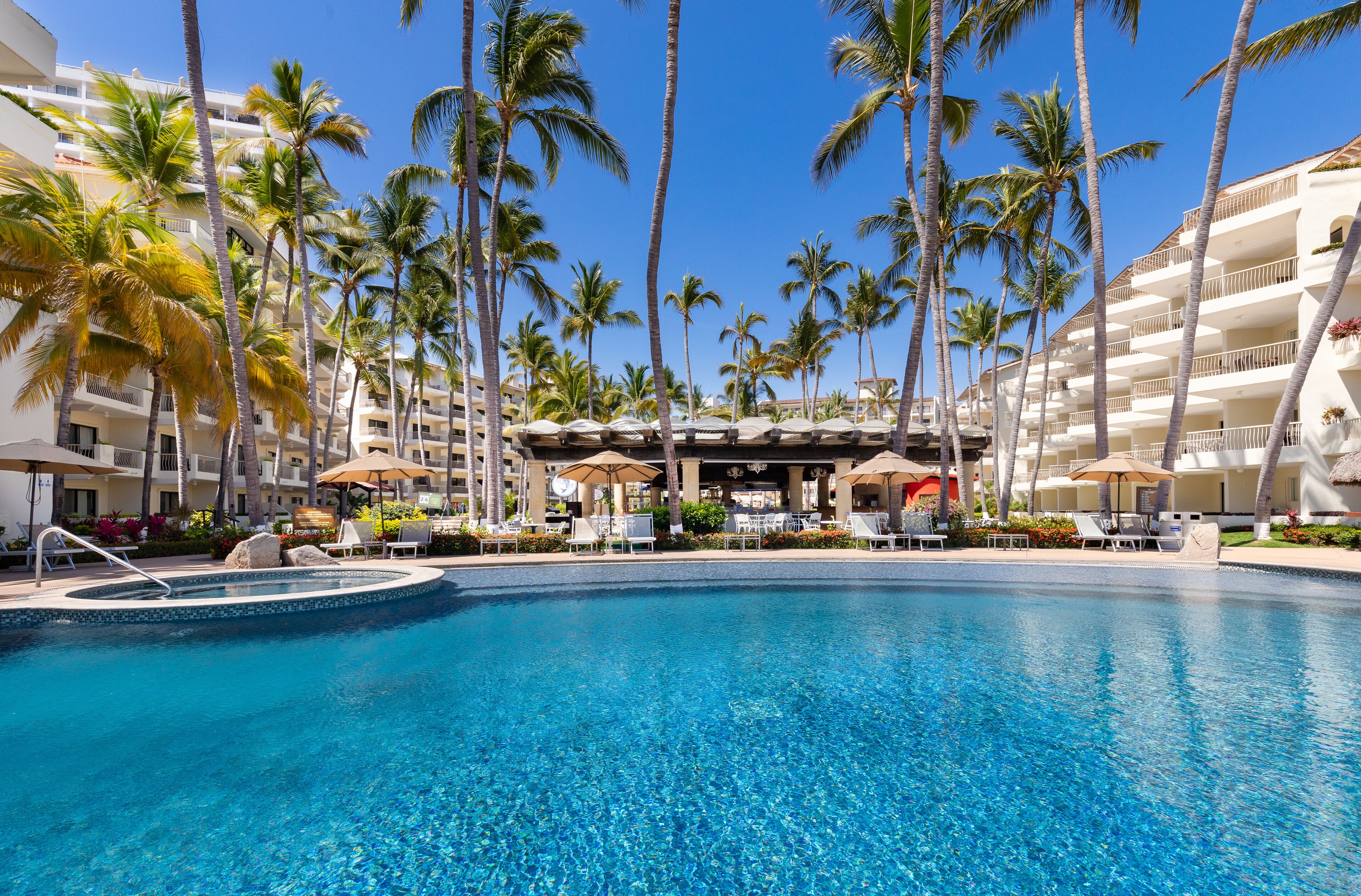A hotel pool with clear blue water, surrounded by palm trees and lounge chairs.