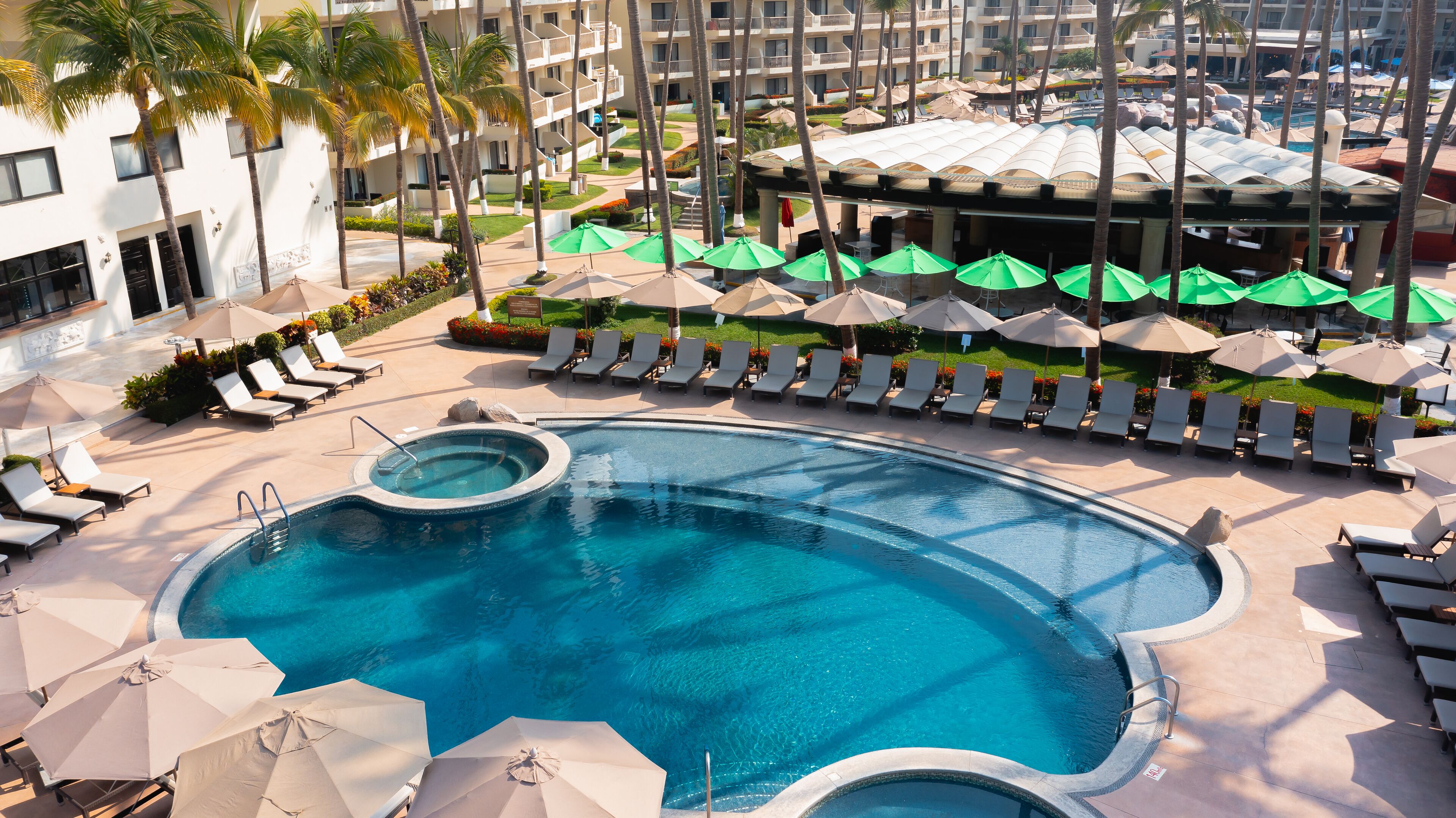 A pool area with multiple lounge chairs and umbrellas, surrounded by palm trees and a building with balconies.