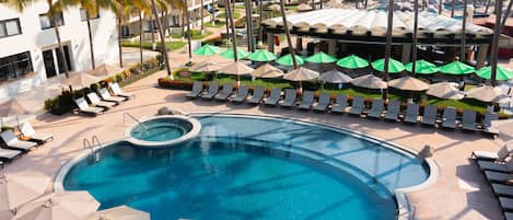 A pool area with multiple lounge chairs and umbrellas, surrounded by palm trees and a building with balconies.