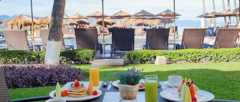 A table set with breakfast, including pancakes, fruit, and a green drink, at a resort with a pool and umbrellas.