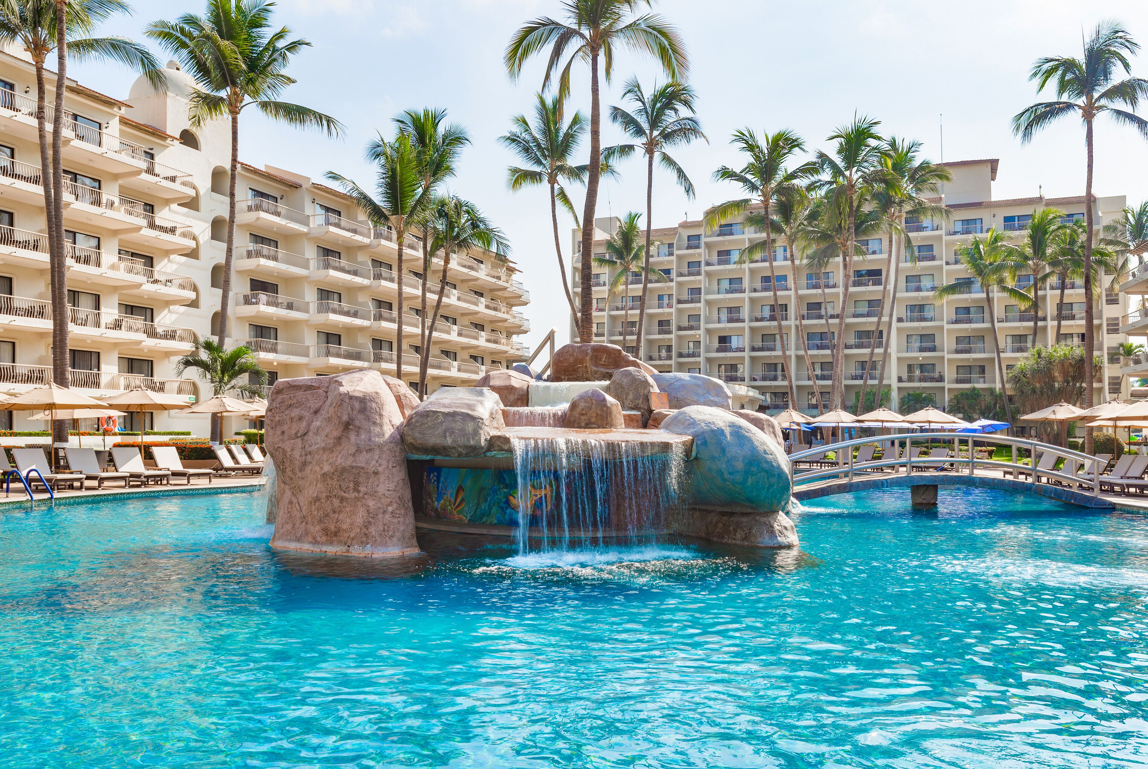 A hotel pool with a waterfall feature, surrounded by palm trees and a multi-story building with balconies.