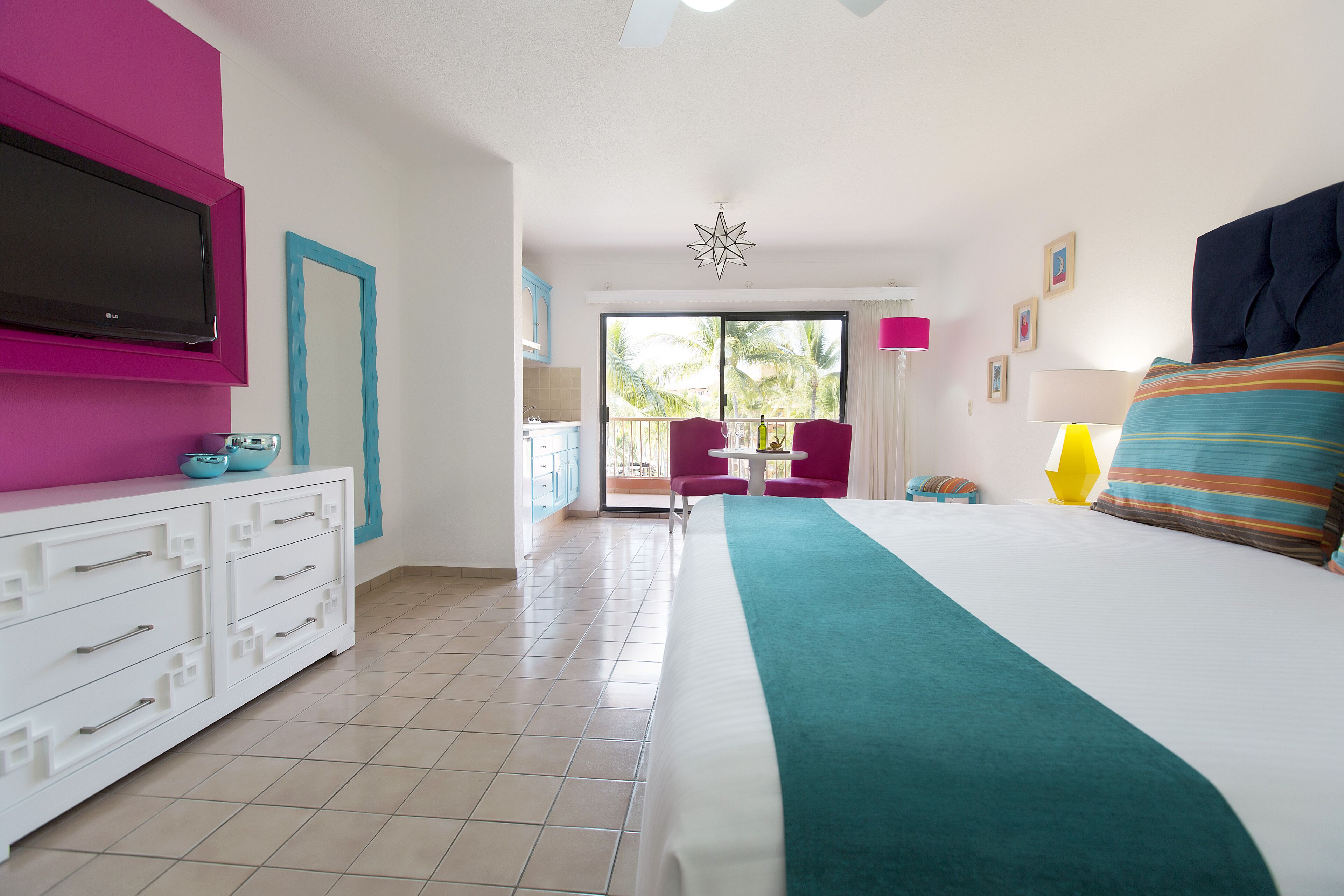 Habitación de hotel con cama, televisión, balcón con una silla y vistas a palmeras.
