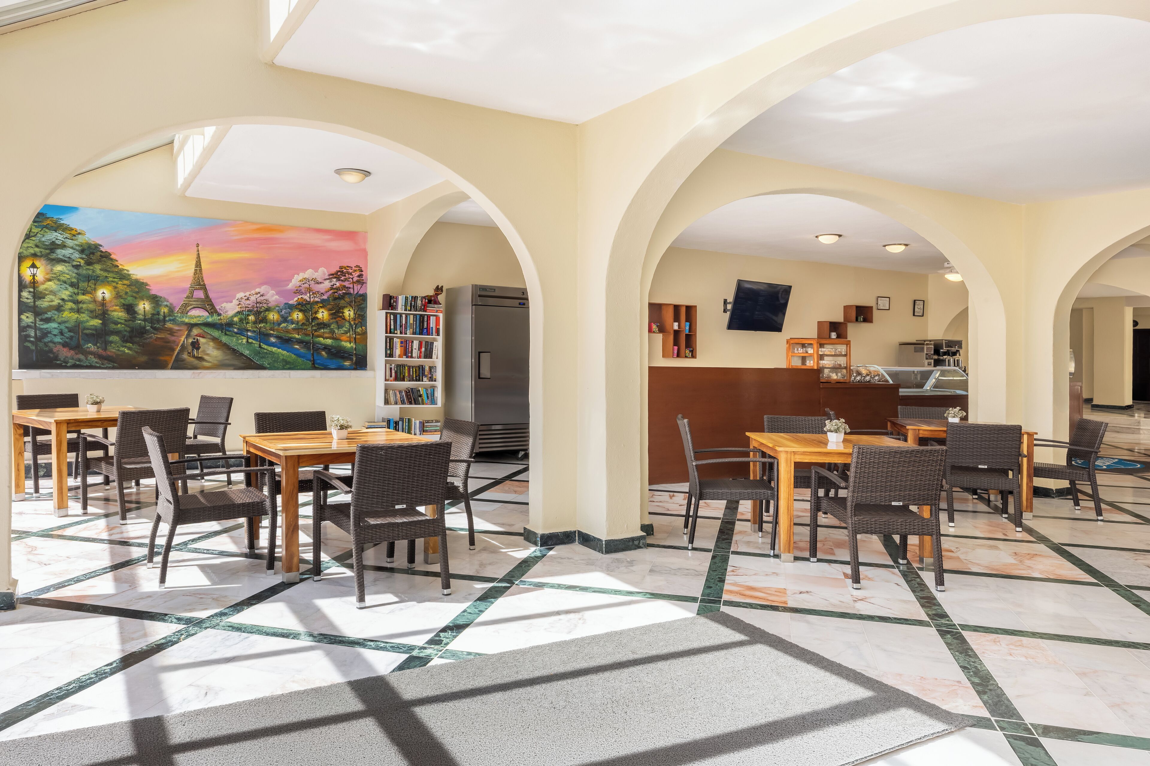 A hotel lobby with a mural of the Eiffel Tower, multiple dining tables with chairs, a refrigerator, and a kitchen area.