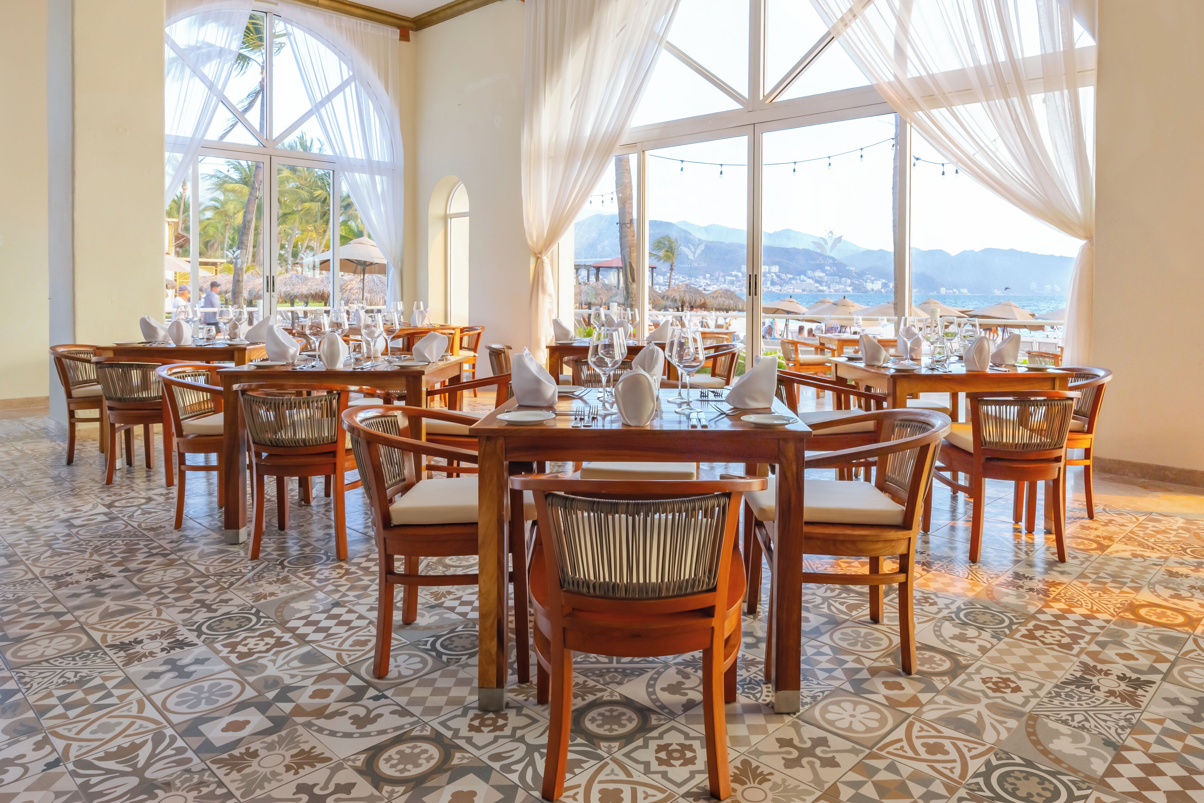 Un ristorante con tavoli e sedie rotonde, ampie finestre con tende e un pavimento decorato.