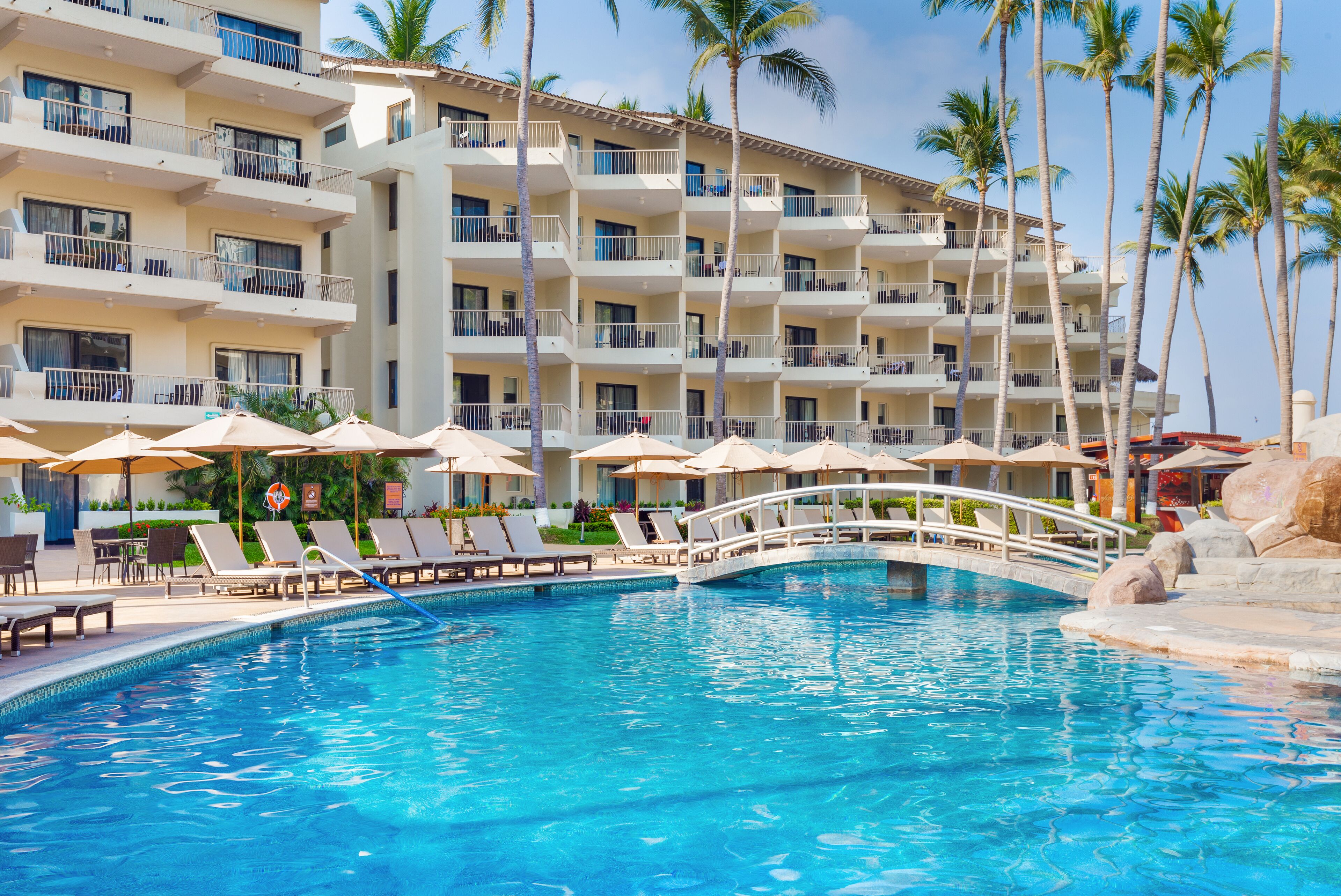 A hotel with a swimming pool, sun loungers, and palm trees.
