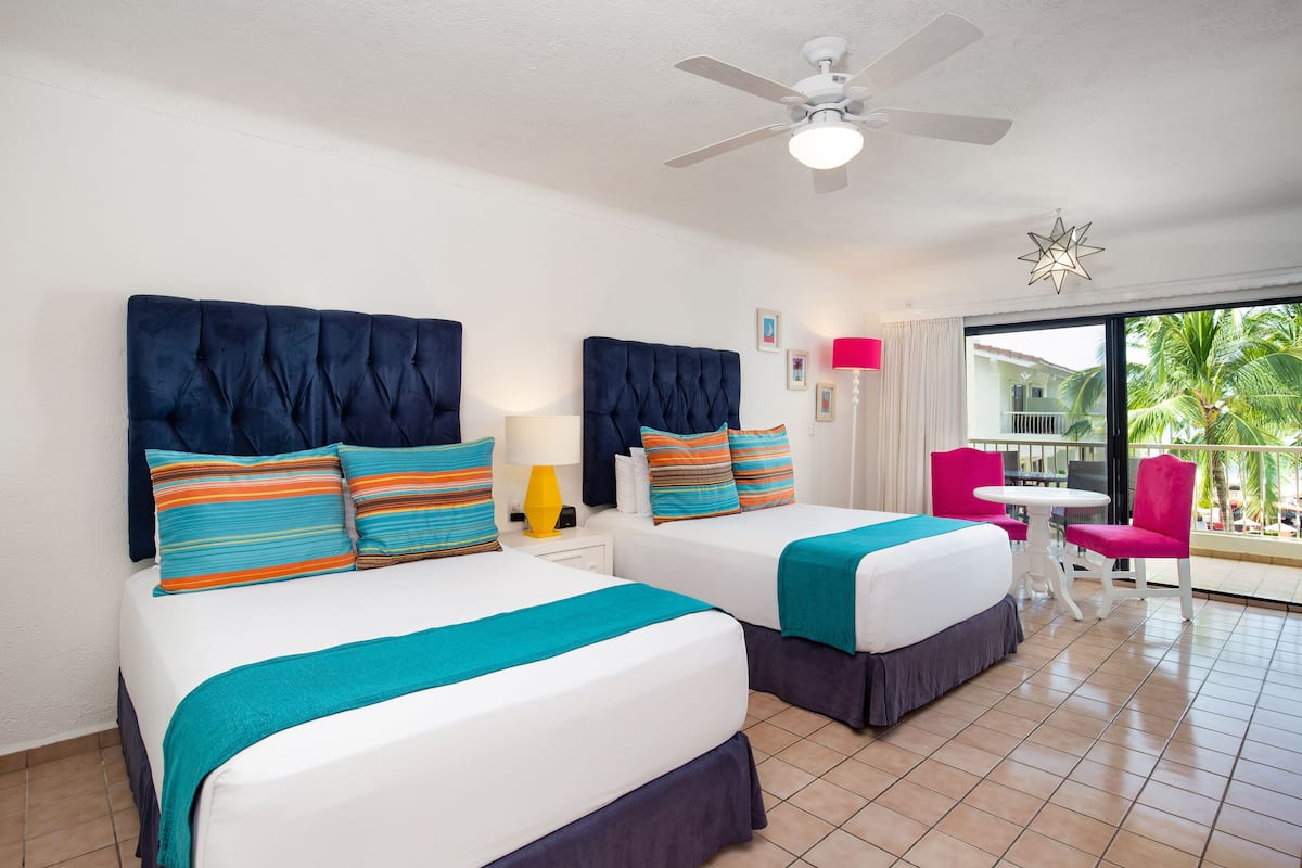 A hotel room with two beds, a ceiling fan, a balcony with a table and chairs, and a view of palm trees.