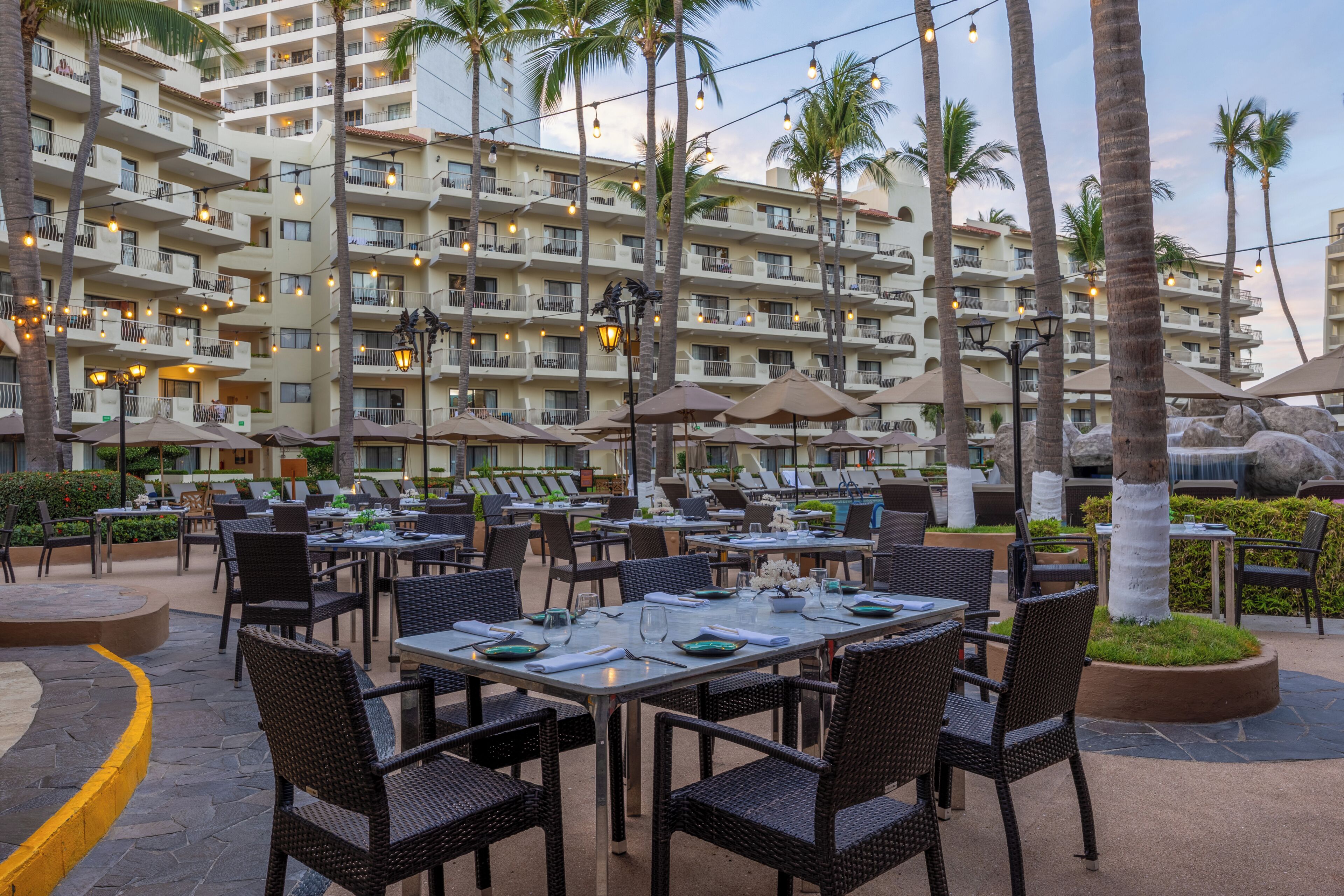 An outdoor dining area with tables and chairs, palm trees, and a multi-story building in the background.