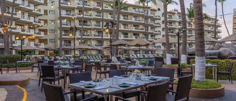 An outdoor dining area with tables and chairs, palm trees, and a multi-story building in the background.