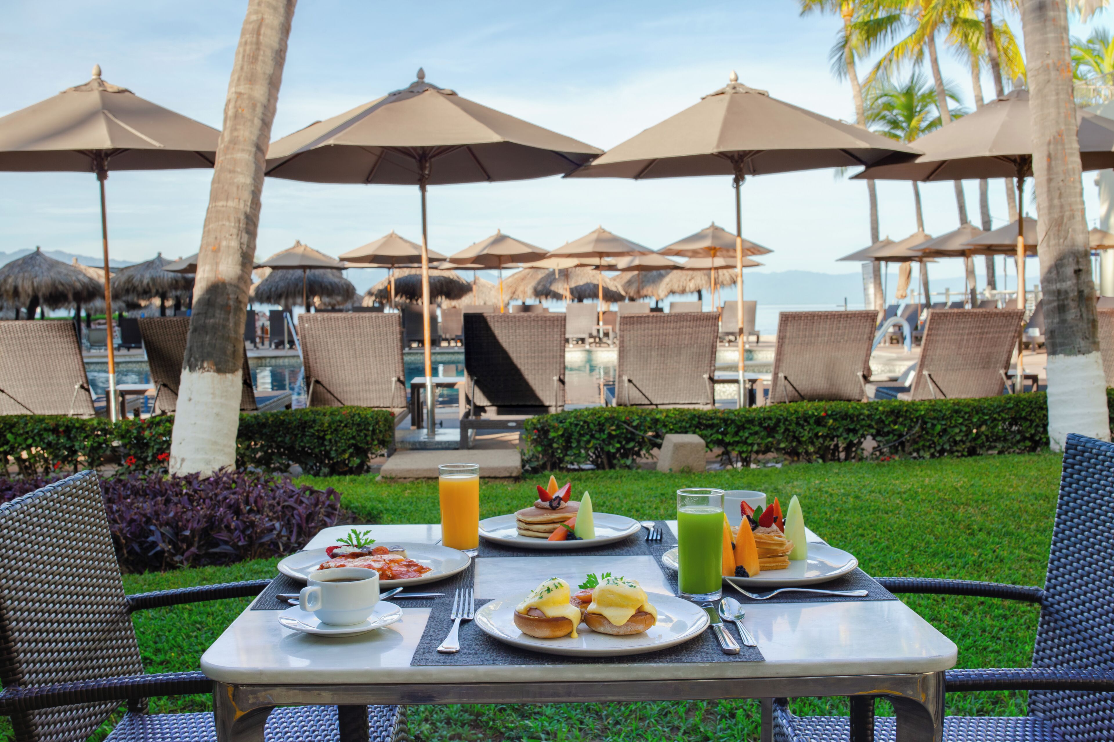 Mesa dispuesta con alimentos para el desayuno, sombrillas exteriores y sillones de descanso en un entorno de resort.