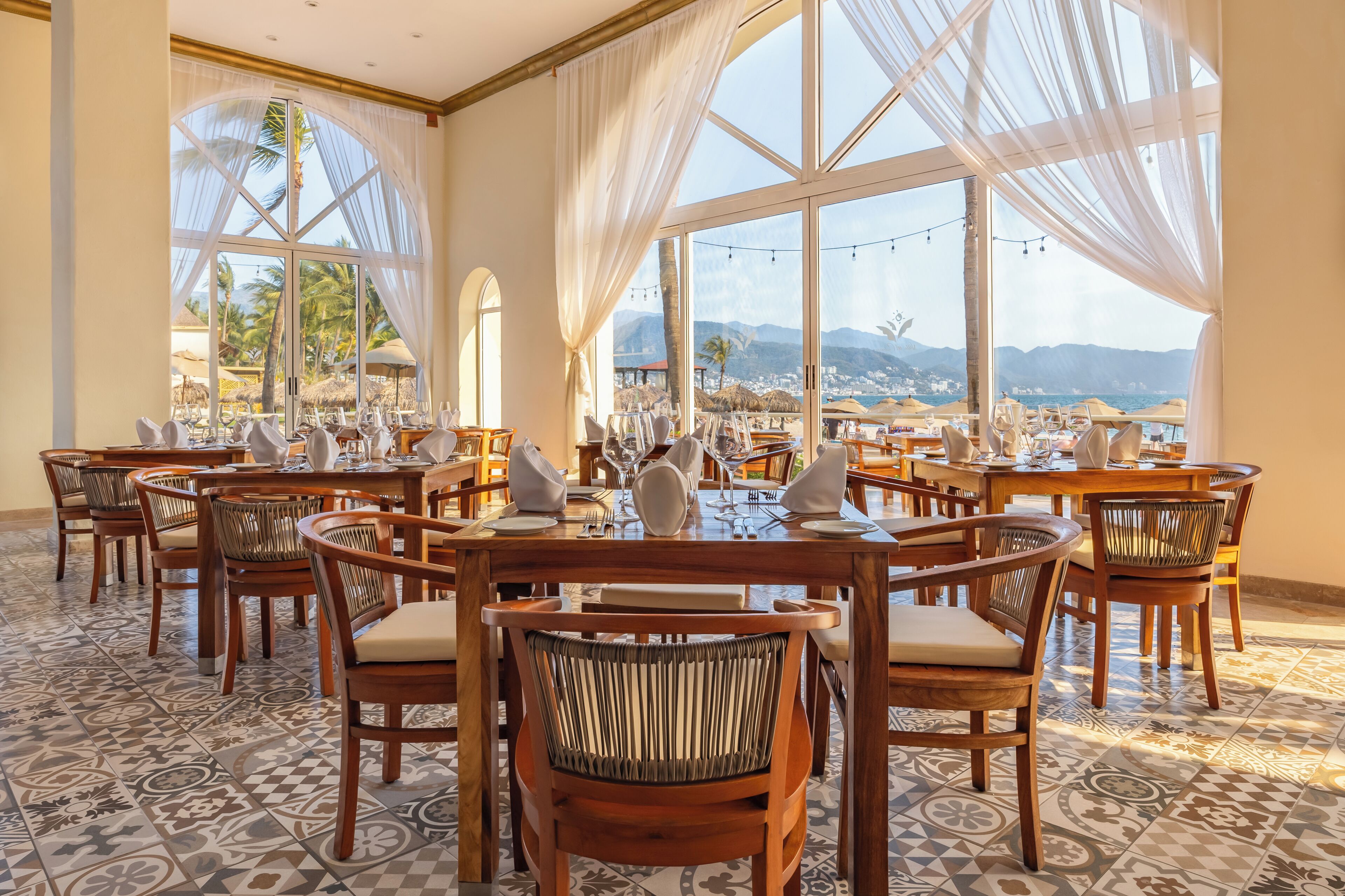 Ein Restaurant mit mehreren Tischen und Stühlen, großen Fenstern mit Vorhängen und Blick auf Strand und Berge.