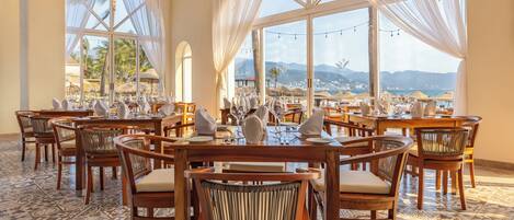 Ein Restaurant mit mehreren Tischen und Stühlen, großen Fenstern mit Vorhängen und Blick auf Strand und Berge.