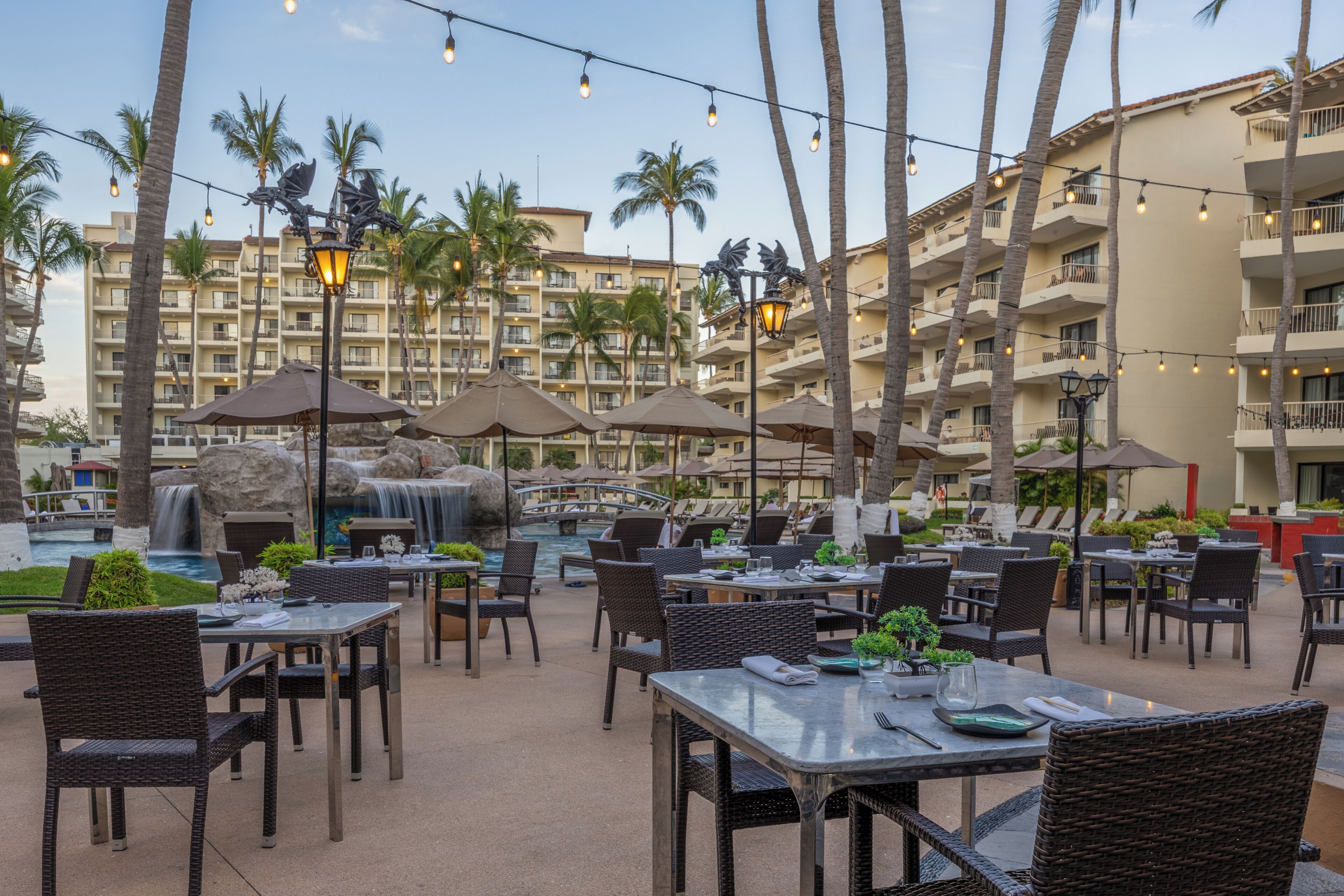 Un espace extérieur aménagé pour les repas, avec des tables et des chaises, des palmiers et un bâtiment en arrière-plan.