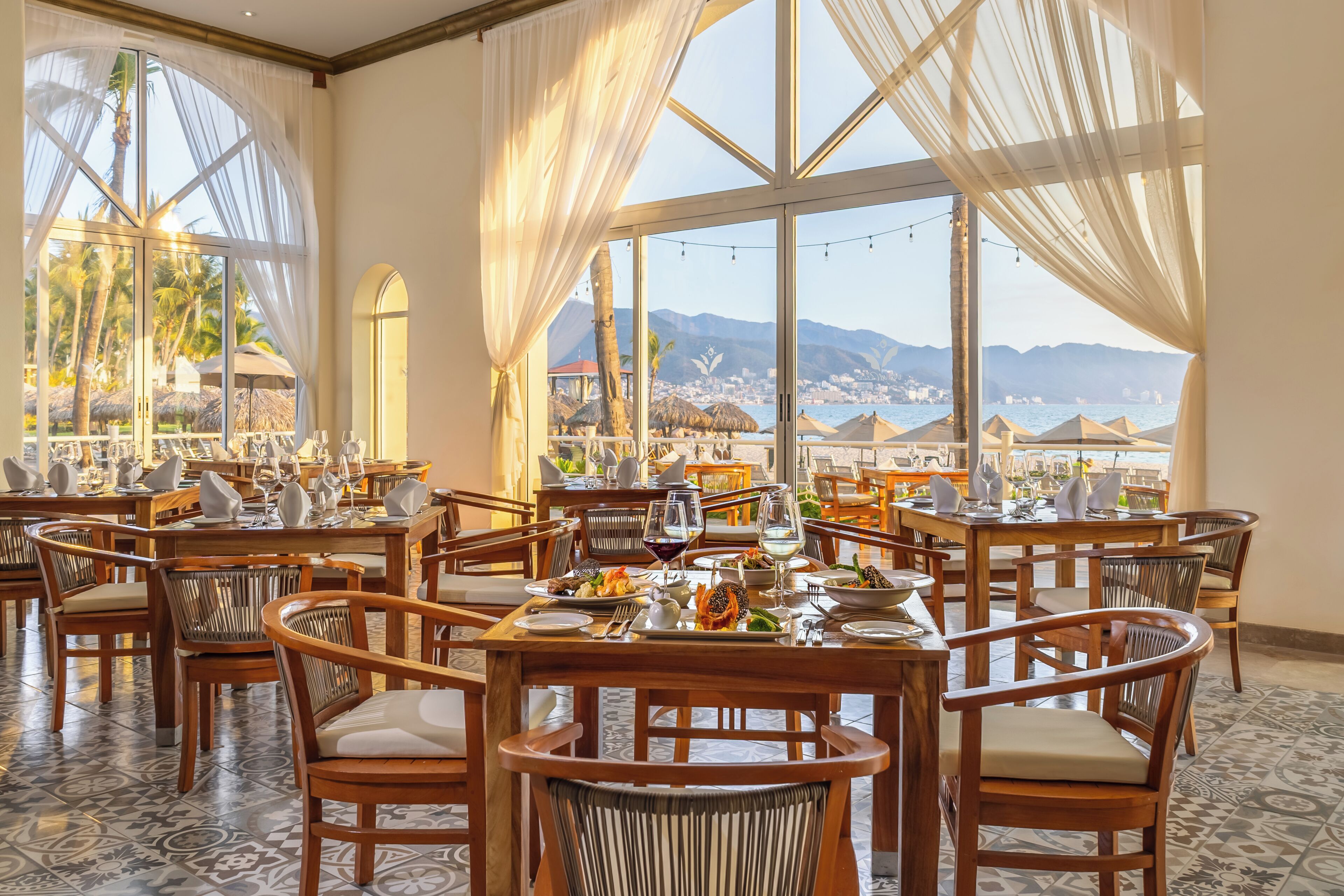 Un restaurante con mesas dispuestas para comer, amplias ventanas con cortinas y vistas al mar y a las montañas.