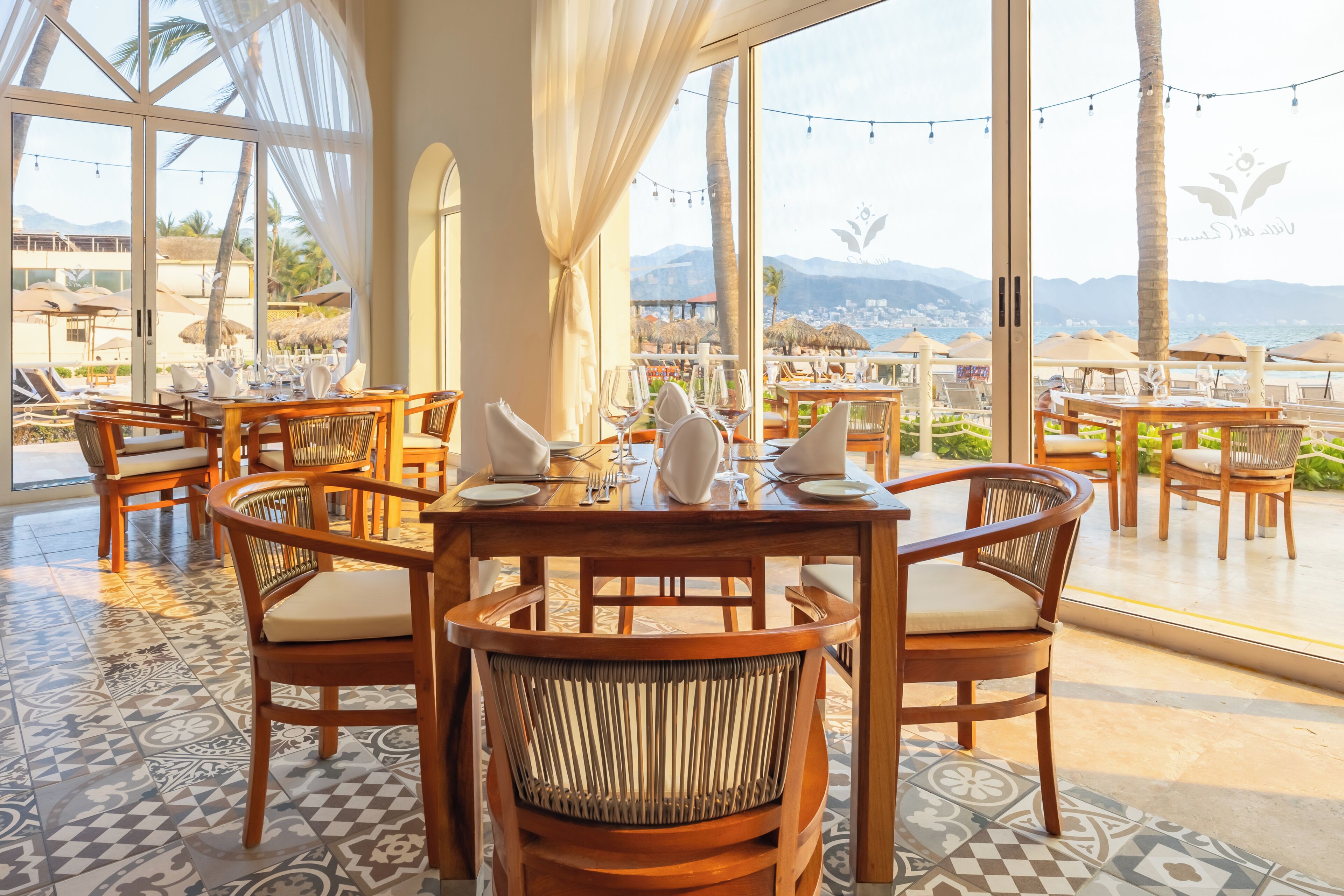 Un restaurante con mesas y sillas de madera, ventanas amplias y vistas a la playa y las montañas.