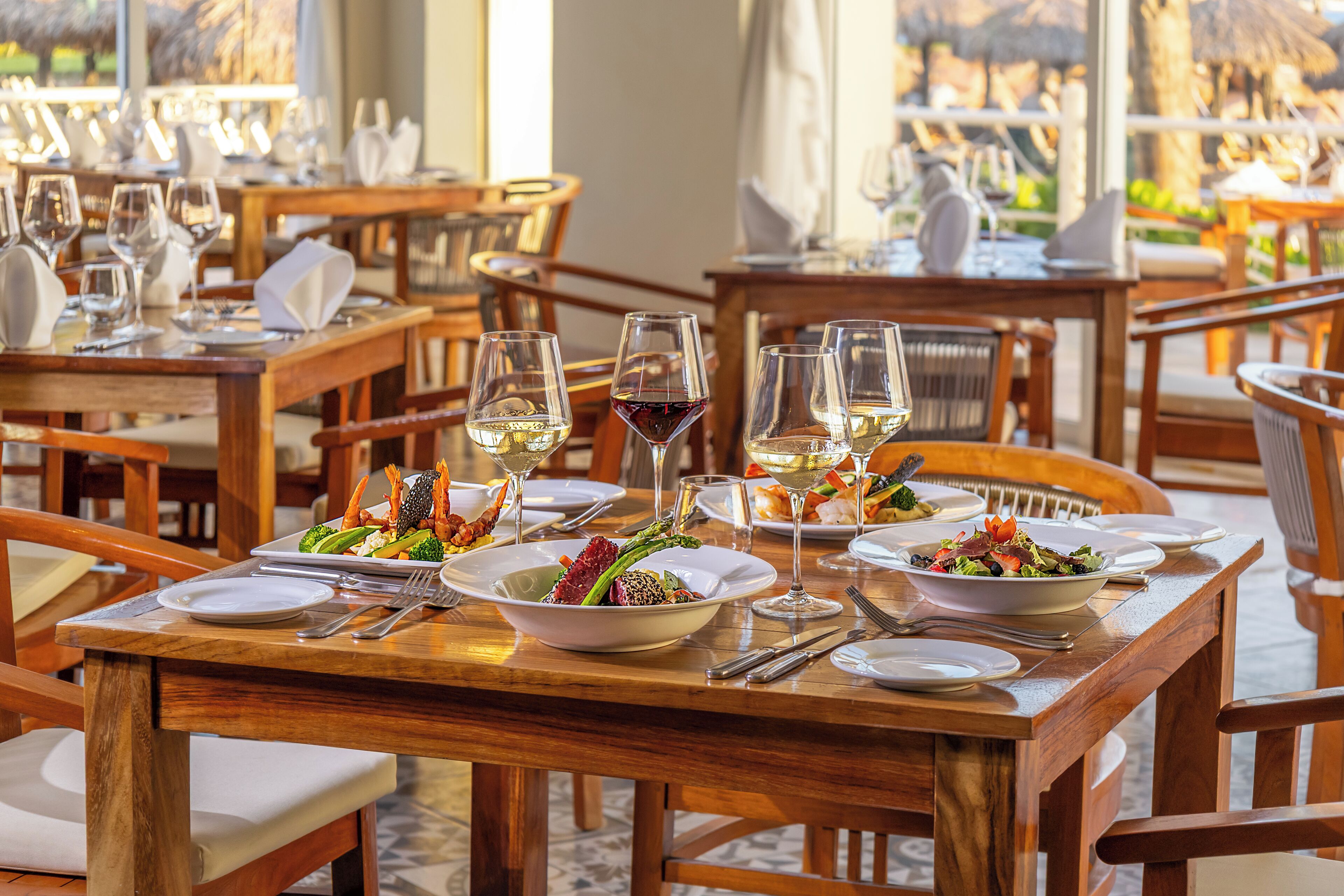 Un ristorante con tavoli in legno, piatti bianchi, bicchieri da vino e vista sull'esterno.