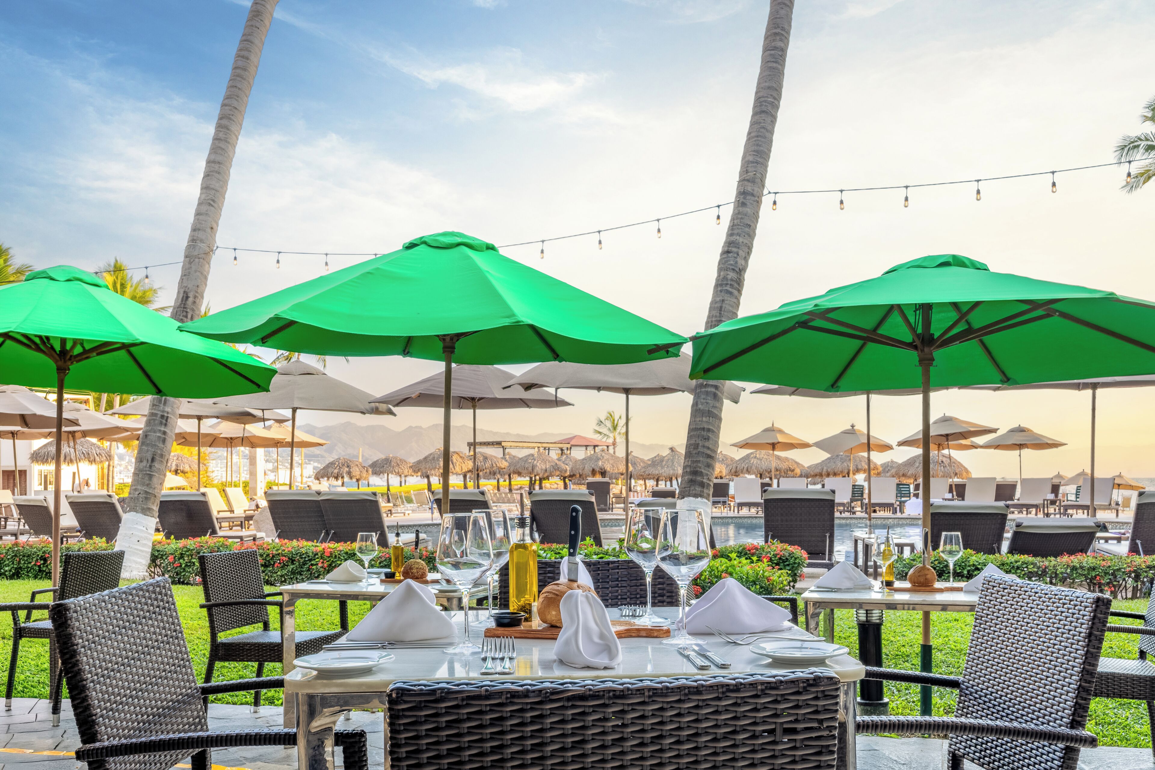 Outdoor dining tables with green umbrellas, wicker chairs, and a view of the pool and palm trees.