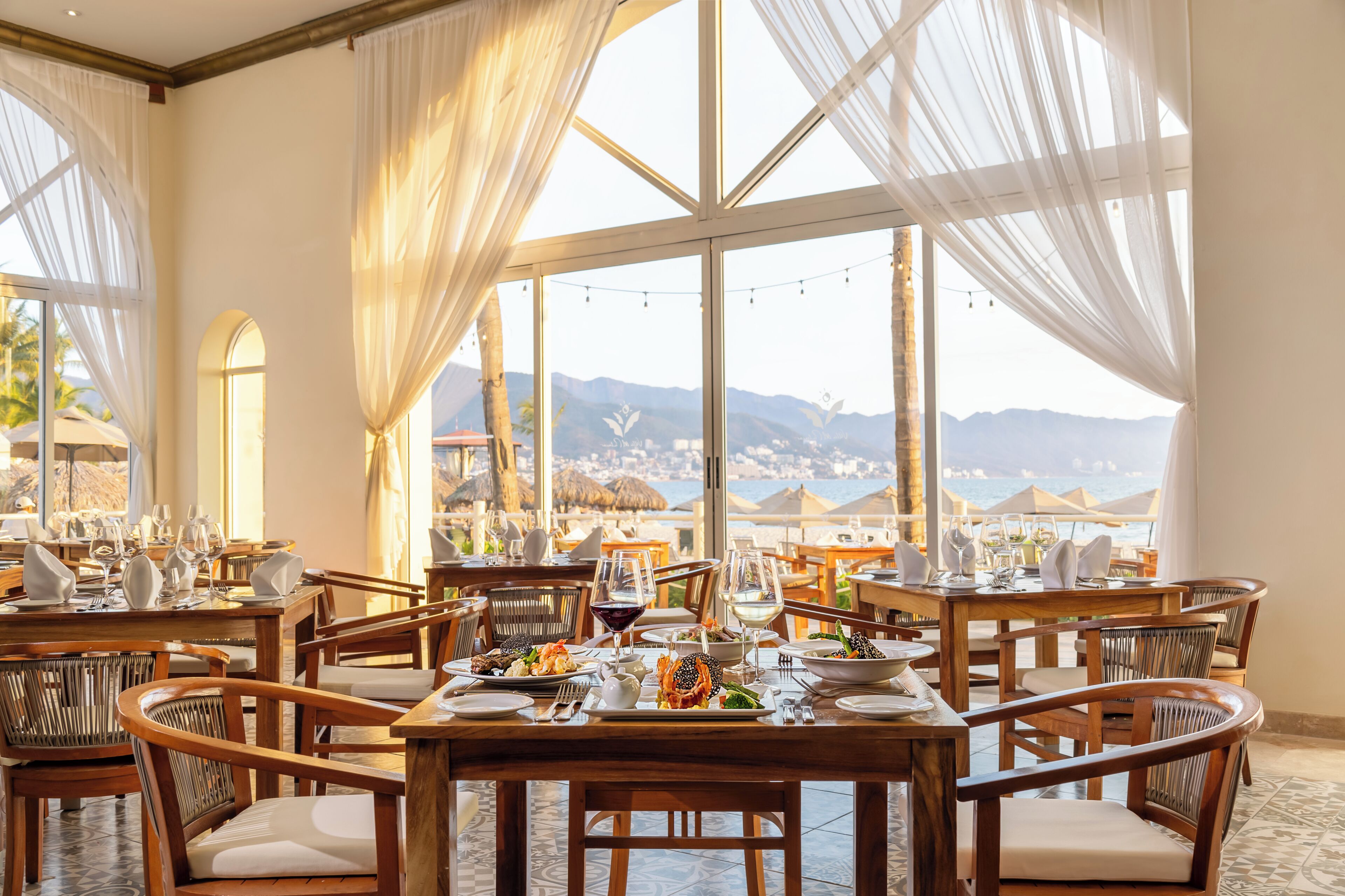 A restaurant with tables set for dining, large windows with curtains, and a view of mountains.