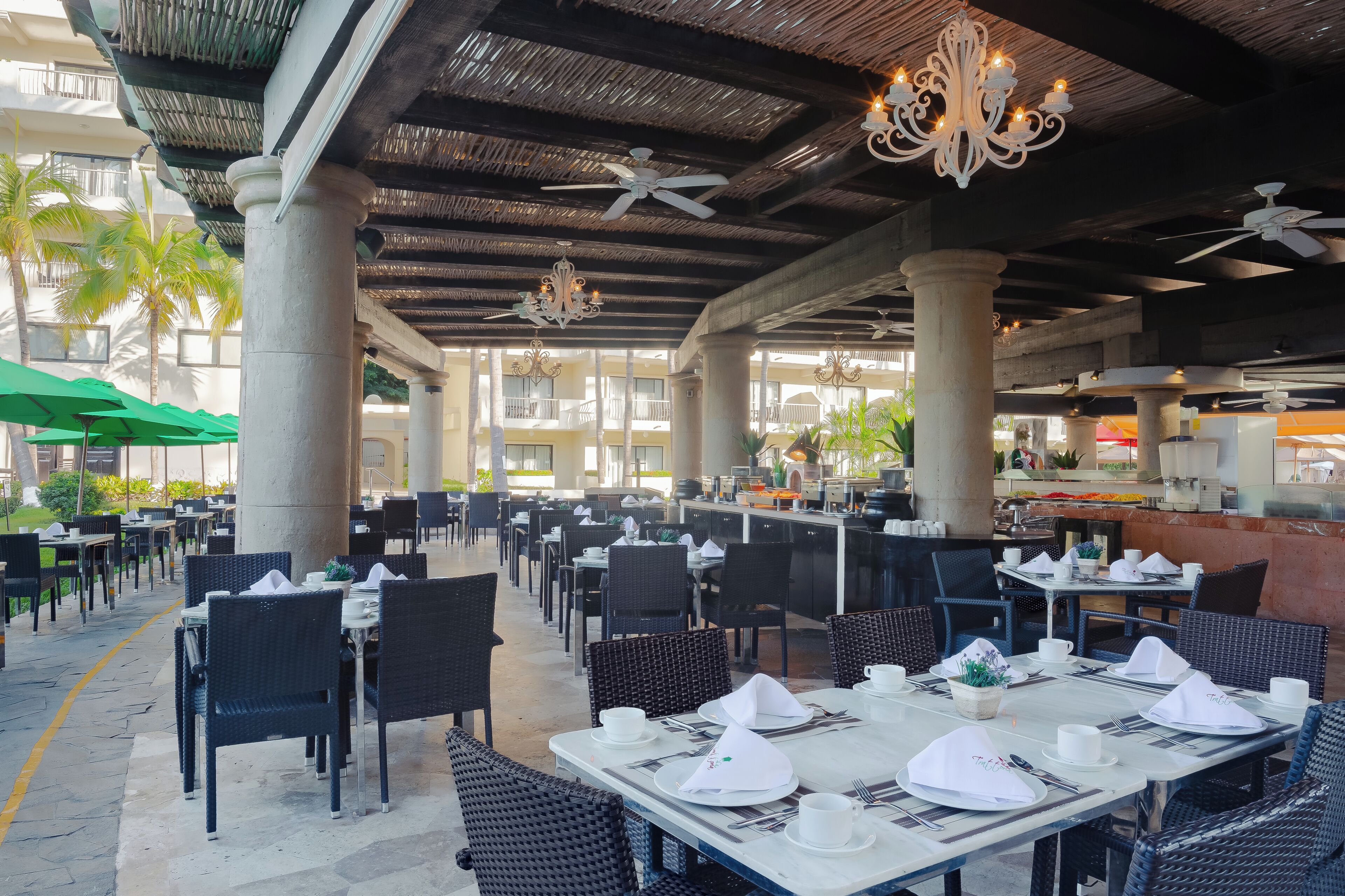 An outdoor dining area with tables set for service, large pillars, ceiling fans, and a bar area.