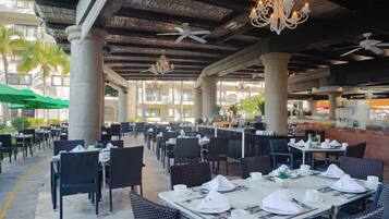 An outdoor dining area with tables set for service, large pillars, ceiling fans, and a bar area.