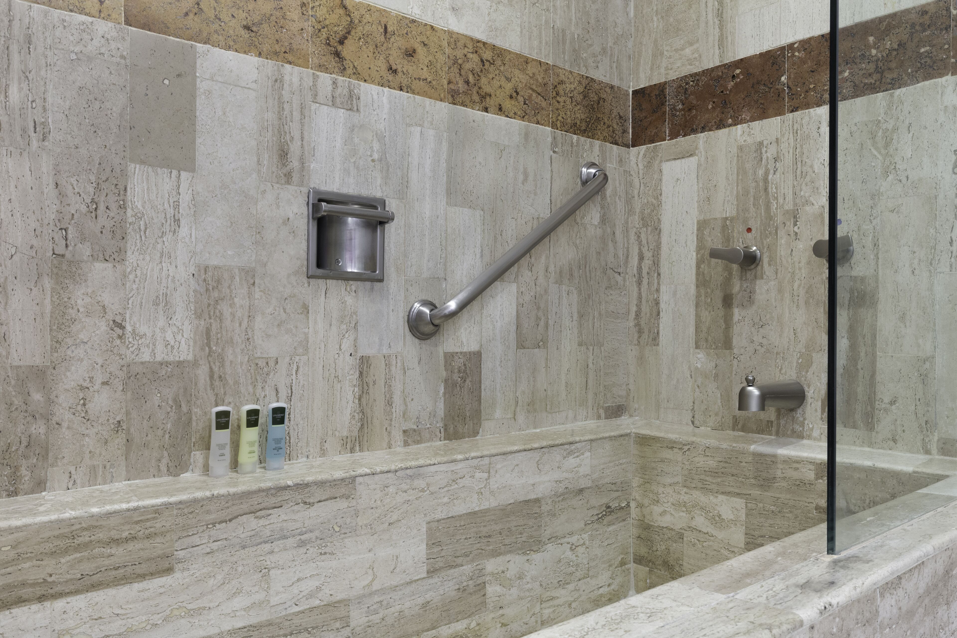 A modern bathroom with a glass shower enclosure, marble tiles, a grab bar, and a shelf with toiletries.
