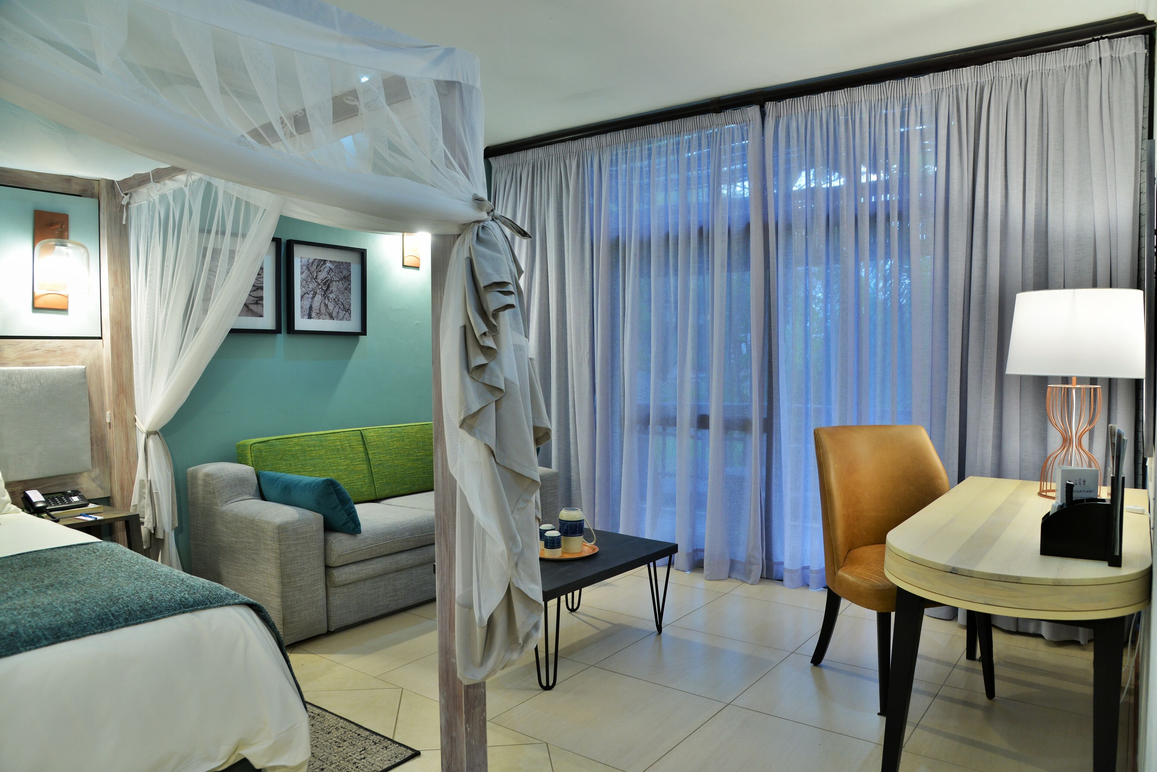 Une chambre d’hôtel avec un lit, un canapé, une table à manger et une chaise. Il y a une fenêtre avec des rideaux, une lampe sur la table et un tableau au mur.