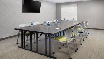 Uma sala de conferências com uma mesa comprida, cadeiras com rodinhas, garrafas de água, uma TV de tela plana e um projetor.