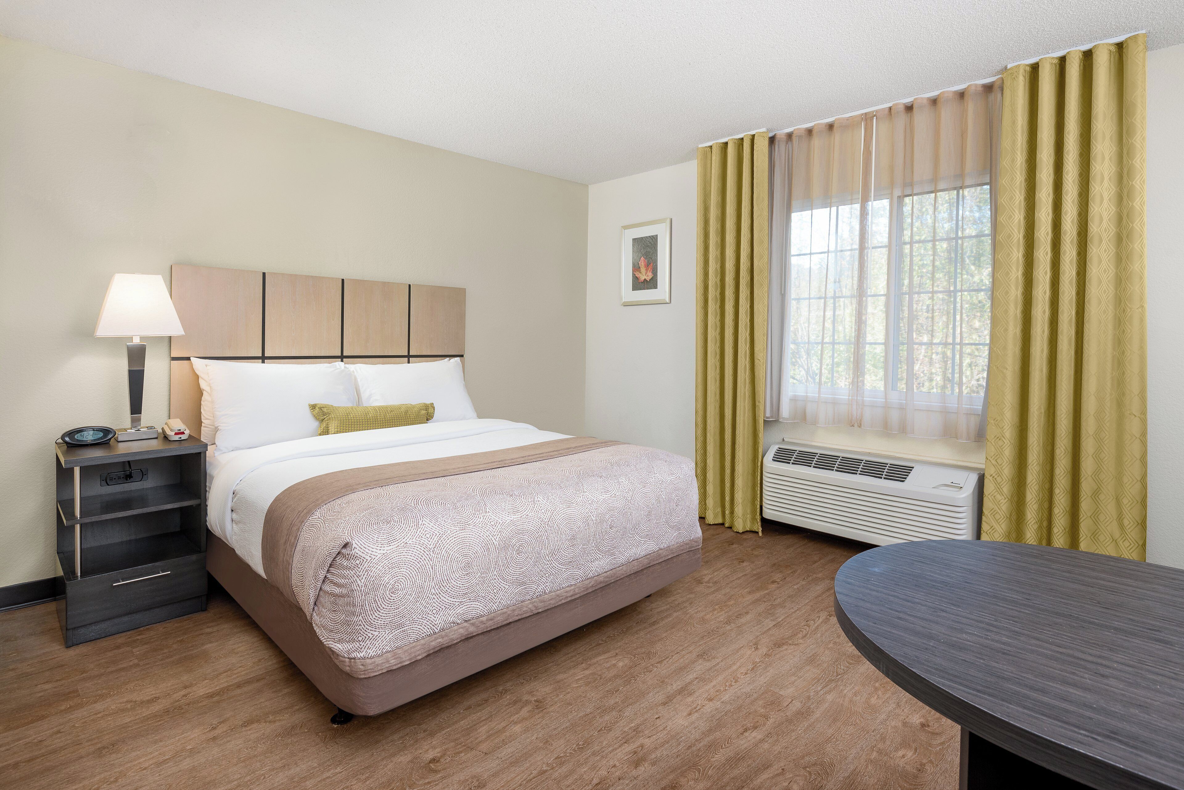 A hotel room with a bed, bedside table, lamp, air conditioning unit, and a window with curtains.