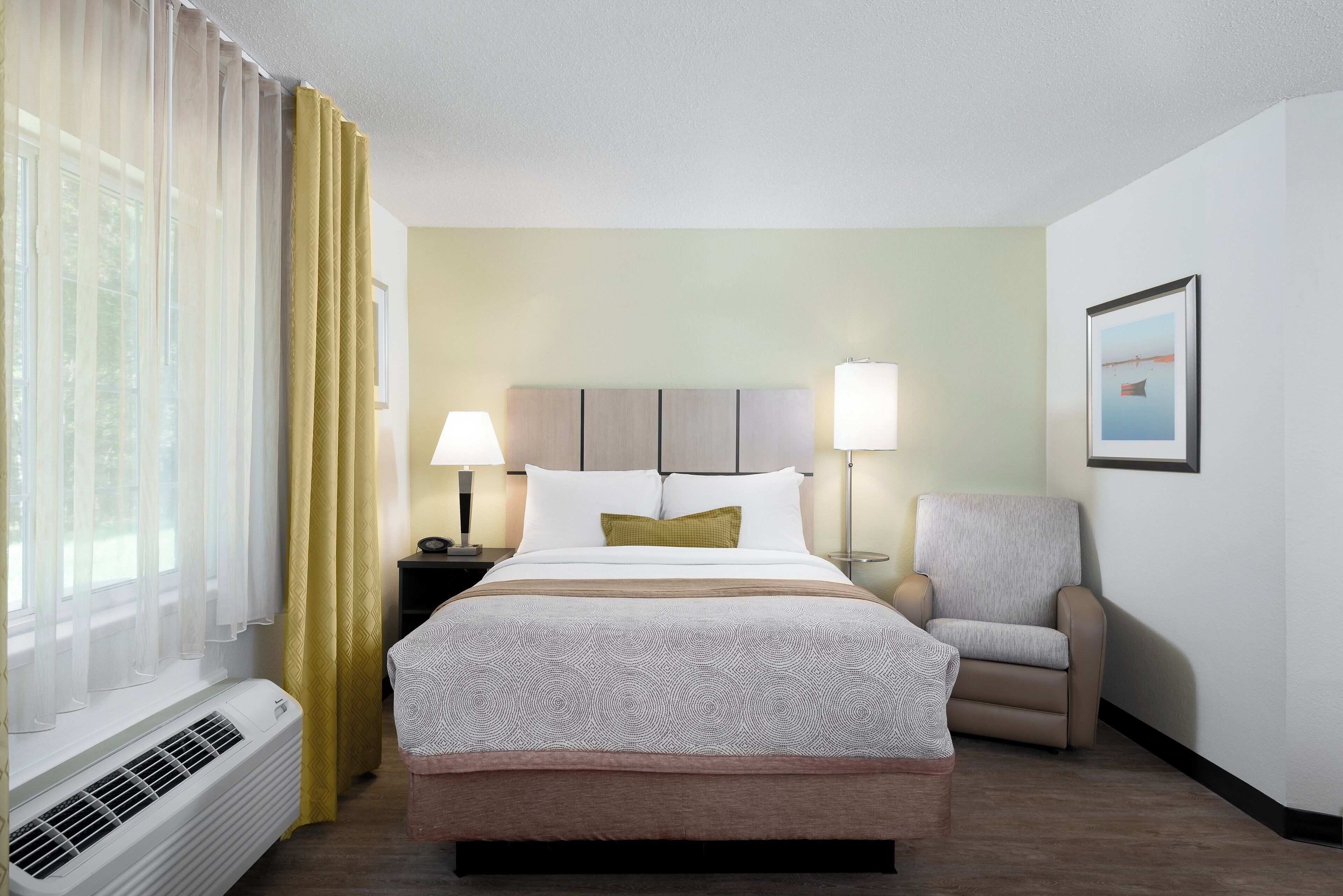 A hotel room with a bed, a chair, a nightstand, a lamp, and a picture on the wall.