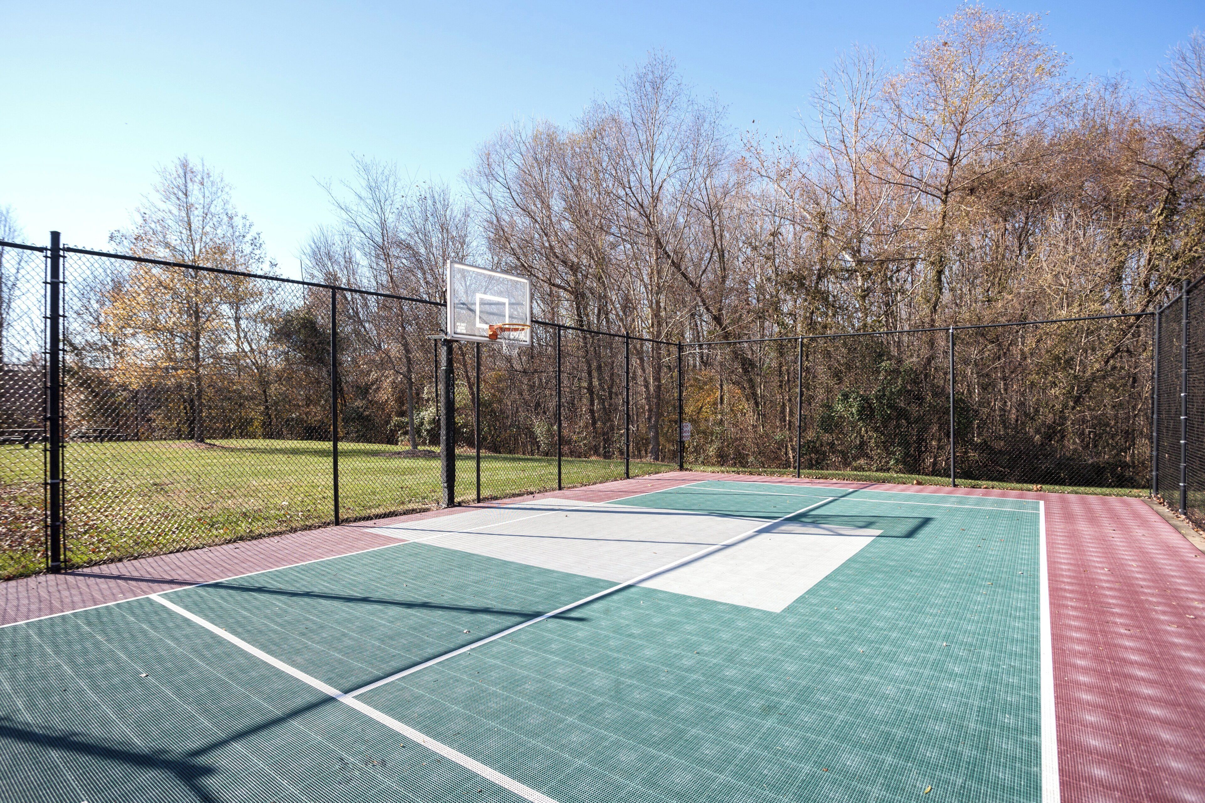 An outdoor basketball court with a chain-link fence, trees, and a clear sky.