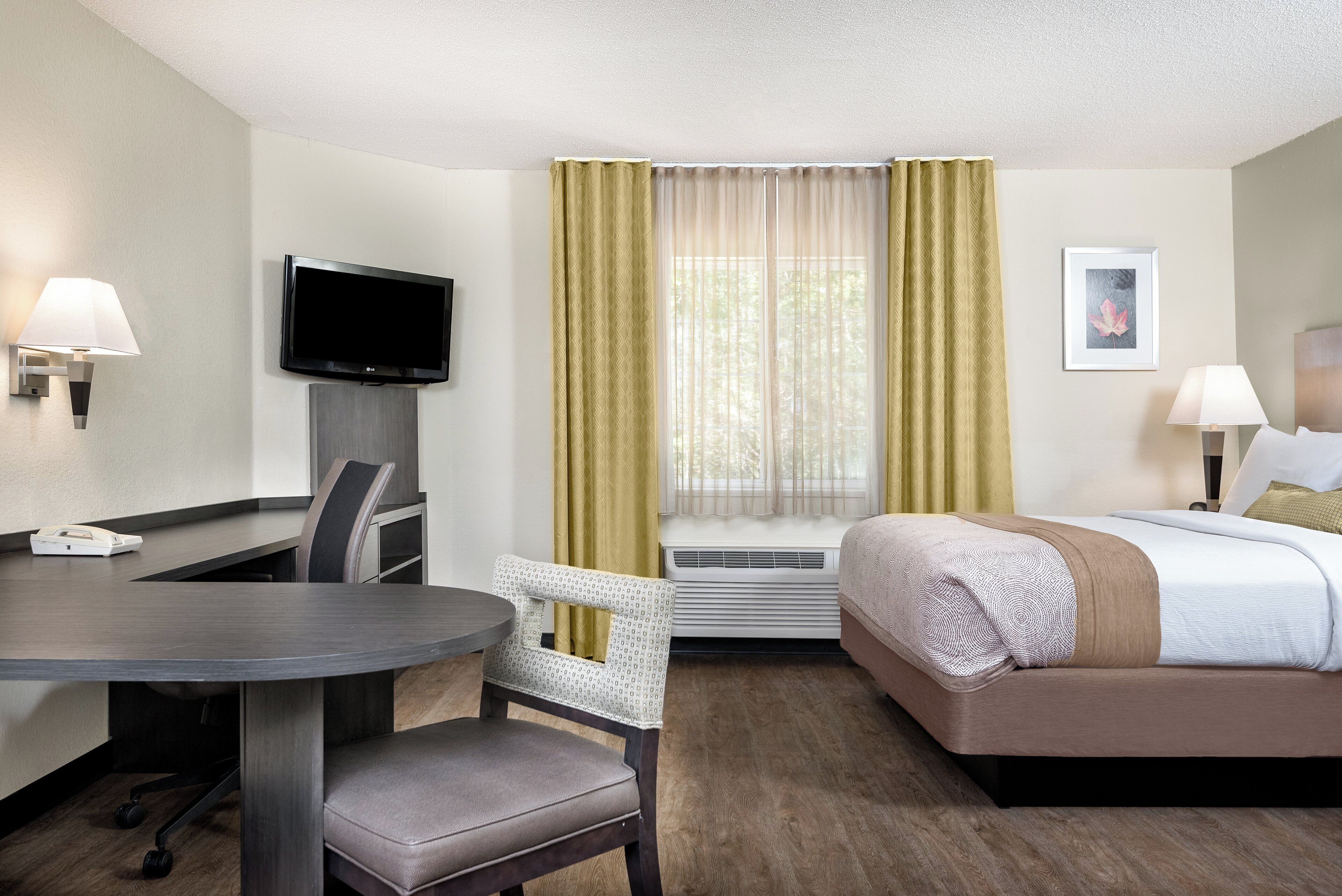 A hotel room with a bed, a desk with a chair, a television, a window with curtains, an air conditioning unit, and a framed picture on the wall.