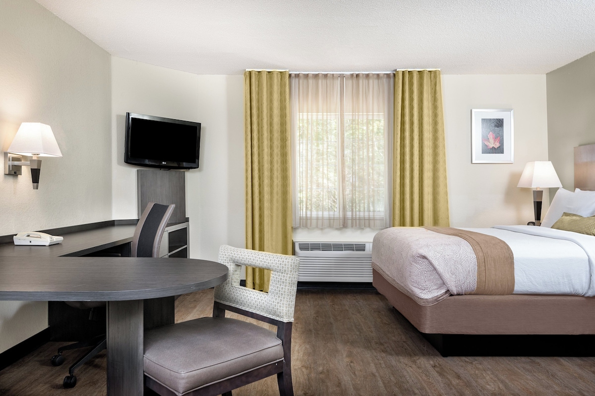 Habitación de hotel con cama, escritorio y silla, televisión, ventana con cortinas, unidad de aire acondicionado y un cuadro en la pared.