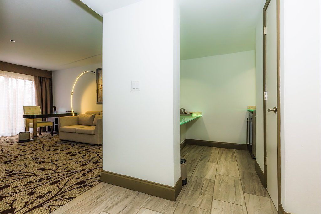 A hotel room with a sofa, a small table, a mirror, a green-lit vanity, and a door leading to another room.