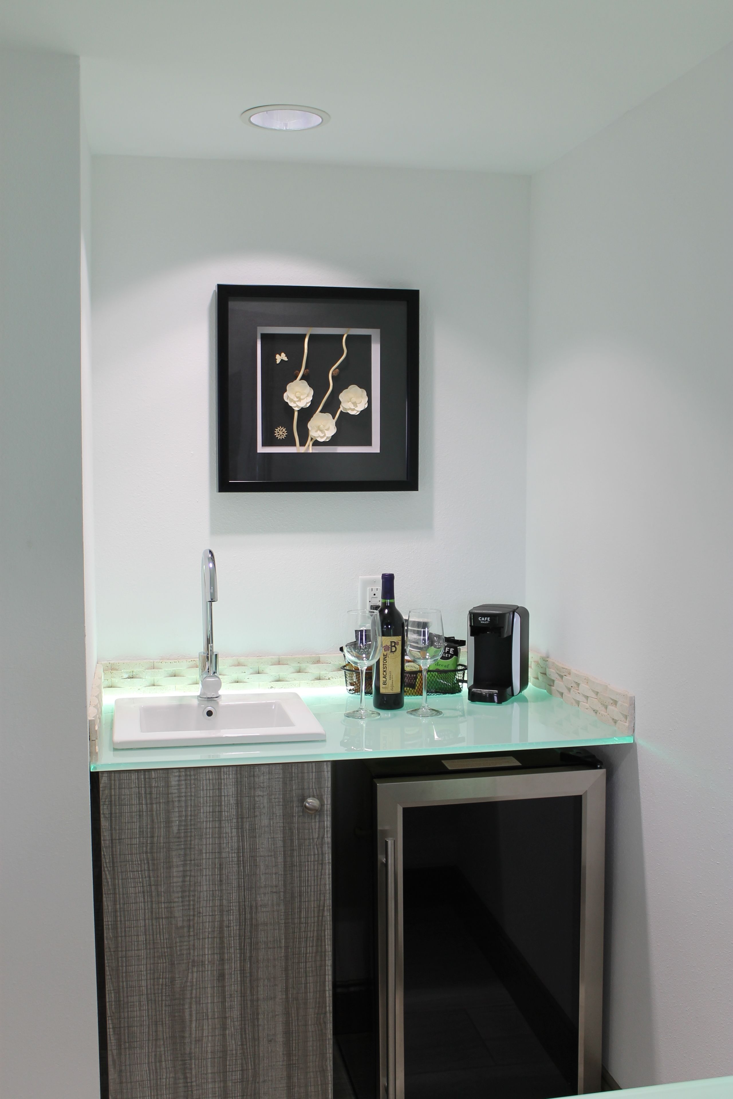 A modern kitchen with a glass countertop, a sink, a framed artwork, and a coffee machine.
