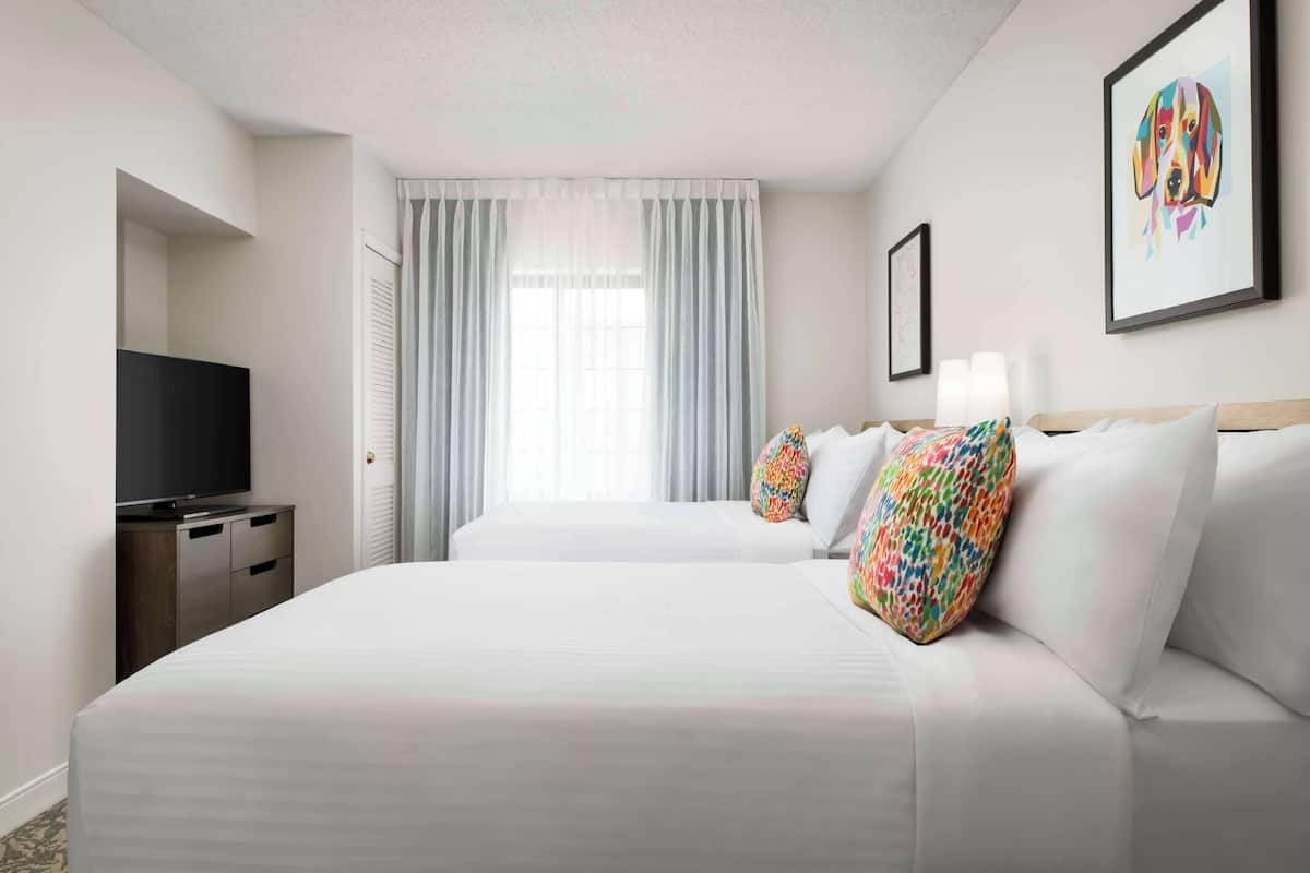 A hotel room with two beds, a television on a dresser, and a large window with curtains.