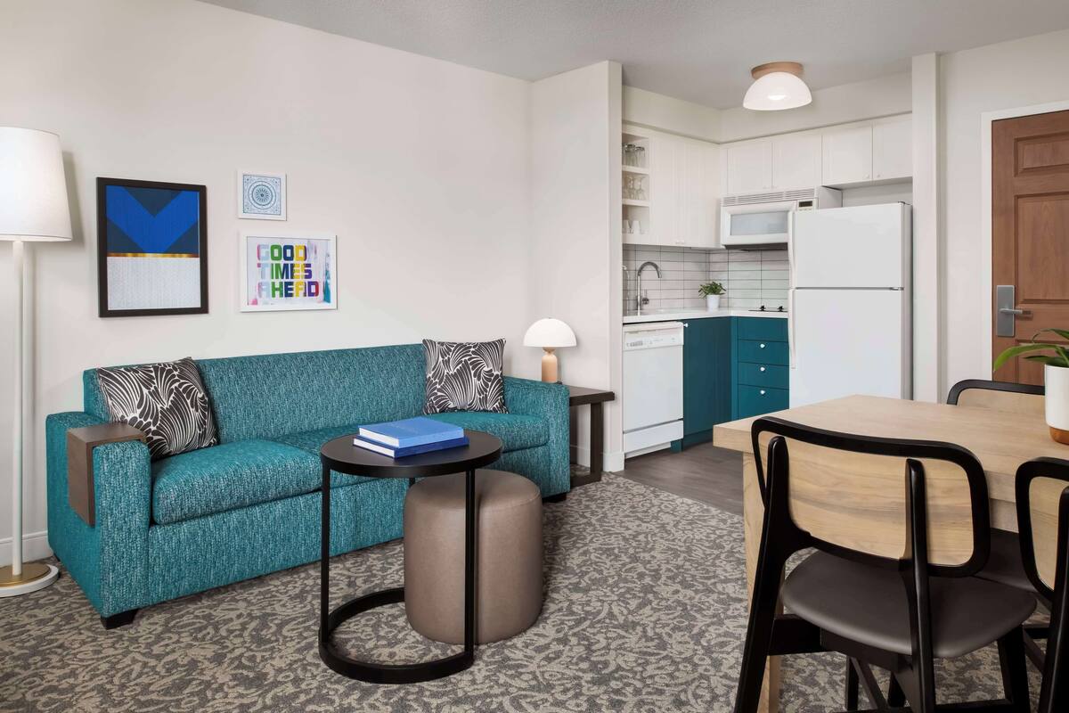 A modern living room with a teal sofa, a small round table, and a dining area with a wooden table and chairs.