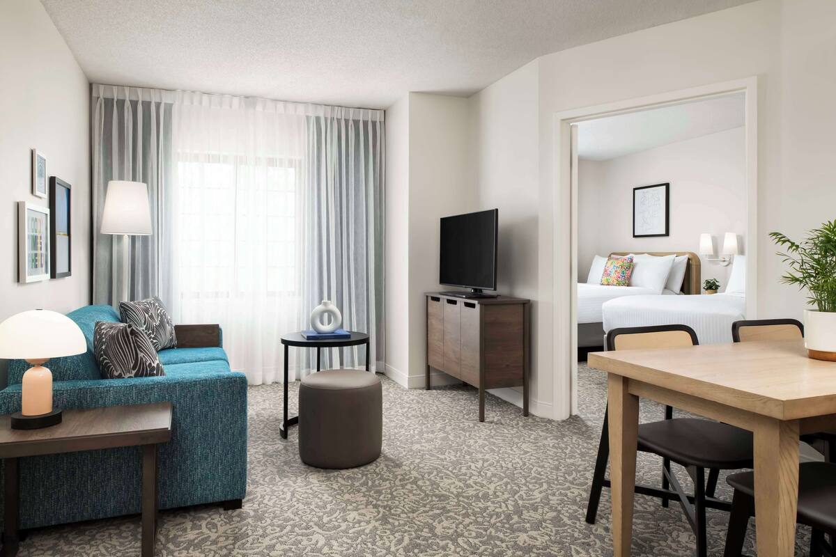 A hotel room with a sofa, ottoman, TV, bedside table, and a dining area with chairs.