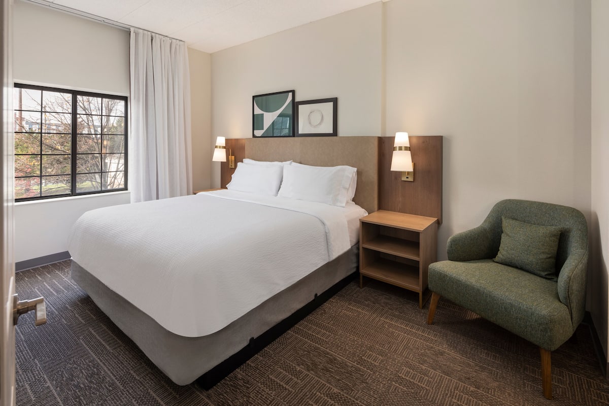 A hotel room with a large bed, a green armchair, a nightstand with lamps, and framed artwork on the wall.