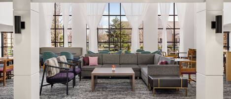 A modern lounge area with a sectional sofa, armchairs, and a coffee table. Large windows with sheer curtains provide natural light and a view of the outdoors.